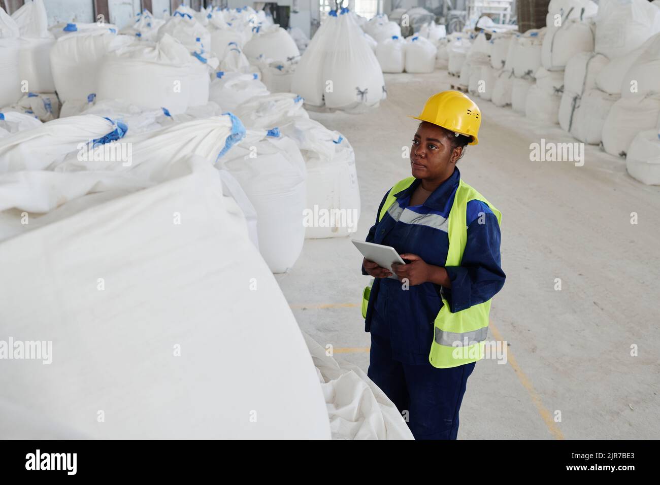Giovane donna nera seria in uniforme e casco di sicurezza usando il tablet mentre in piedi di fronte a sacco bianco enorme con materie prime Foto Stock