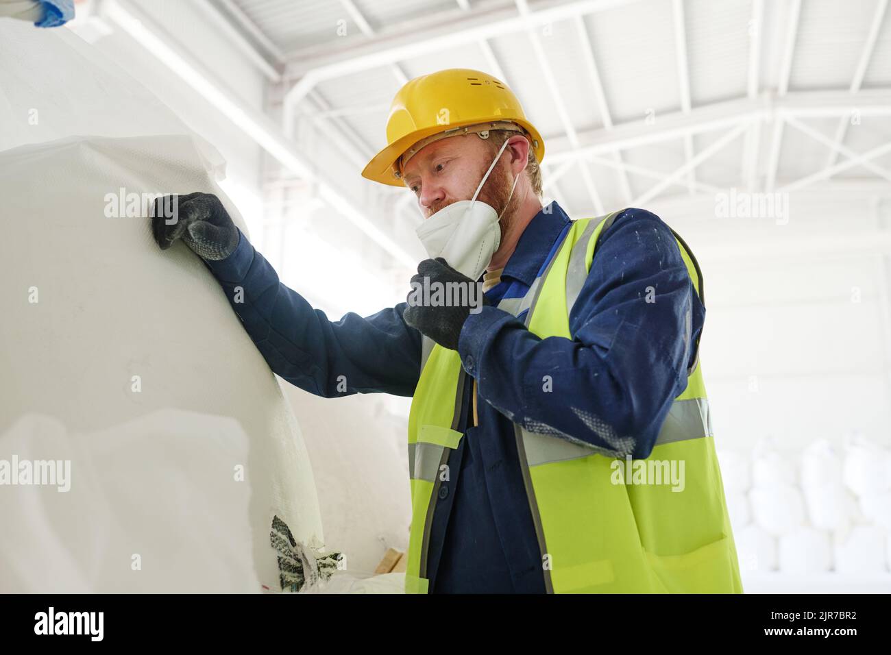 Ingegnere di fabbrica moderna o impianto di produzione mettendo respiratore sul suo viso mentre si trova di fronte a enorme sacco bianco con materie prime Foto Stock