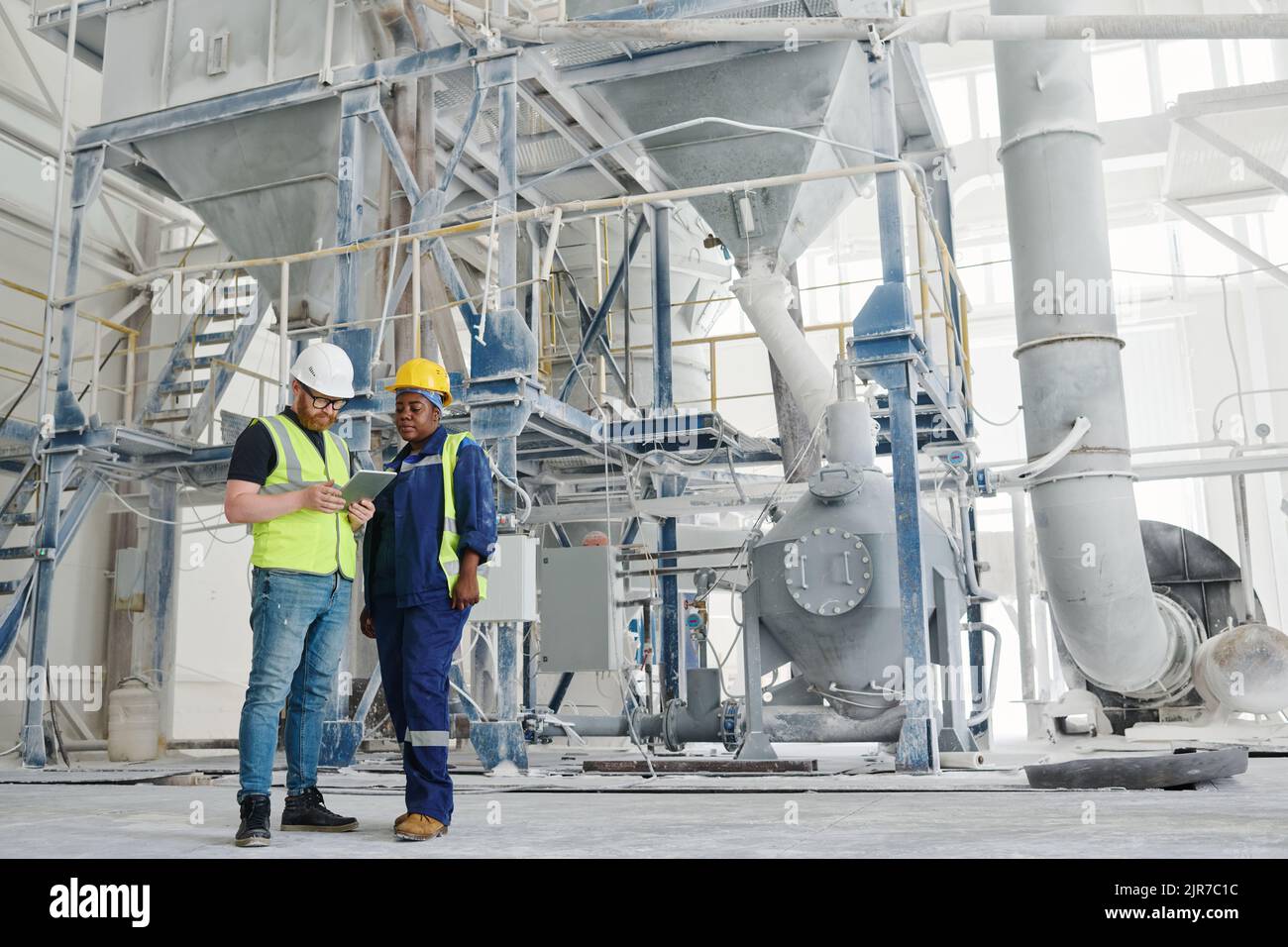 Due giovani colleghi multiculturali nel settore dell'abbigliamento da lavoro e degli hardhat che guardano la guida manuale online durante il lavoro in fabbrica moderna Foto Stock