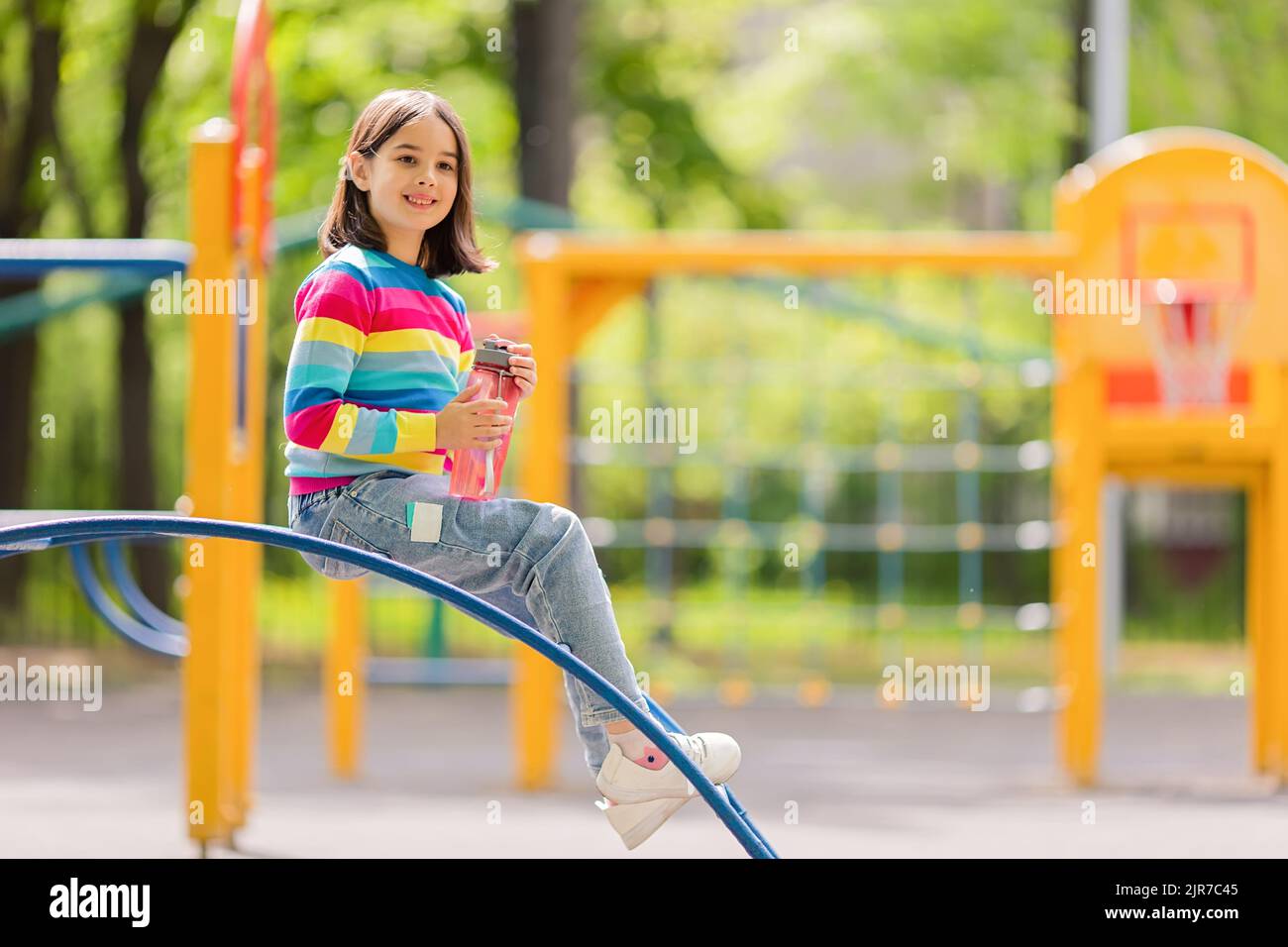 Ragazza sorridente carina, 5-6 anni, seduta con una bottiglia d'acqua di plastica nel parco giochi Foto Stock