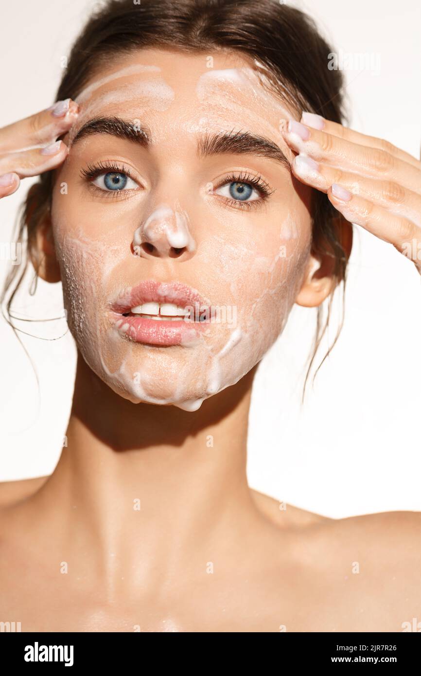 Ritratto verticale di giovane donna lavando il viso con gel di schiuma detergente, sorridendo felice, pulendo la sua pelle facciale, in piedi su sfondo bianco Foto Stock