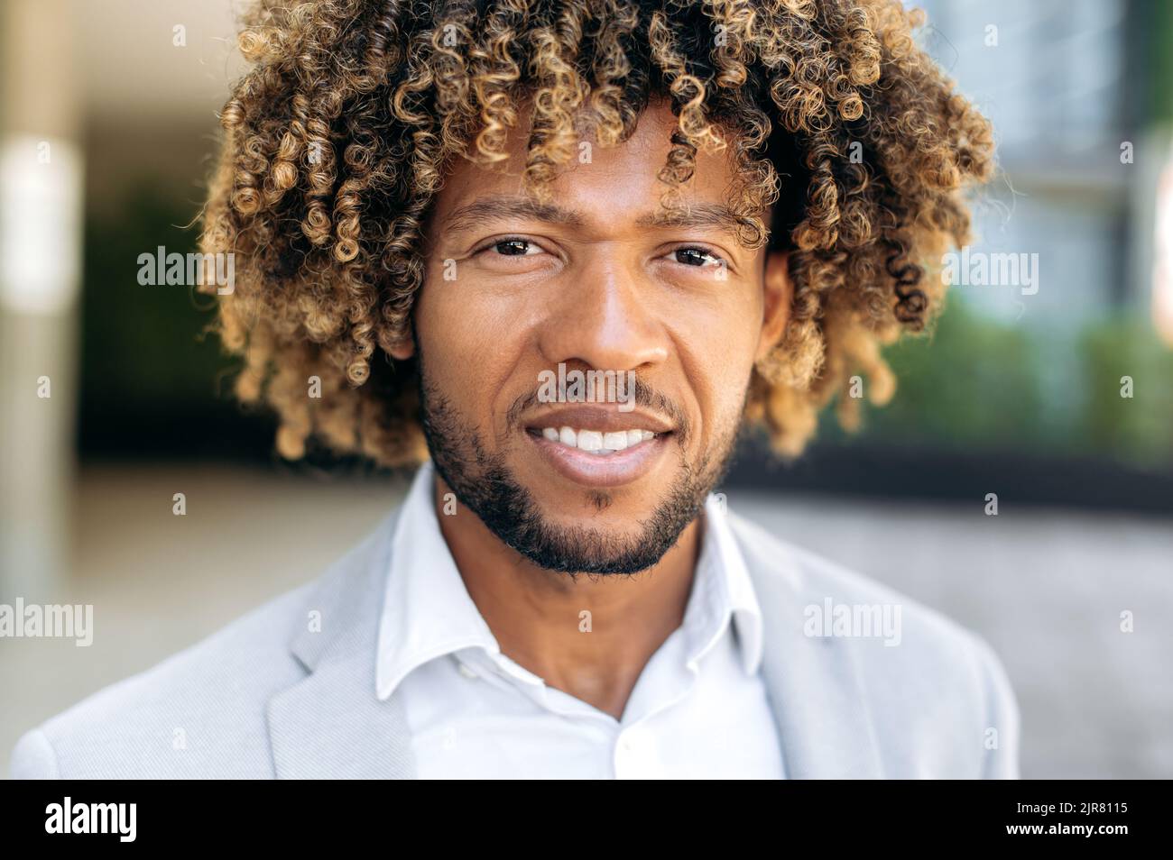 Foto ravvicinata di un bel carismatico uomo brasiliano o ispanico dai capelli ricci fiducioso, imprenditore di successo, in piedi all'aperto, guardando la fotocamera, sorridente amichevole Foto Stock