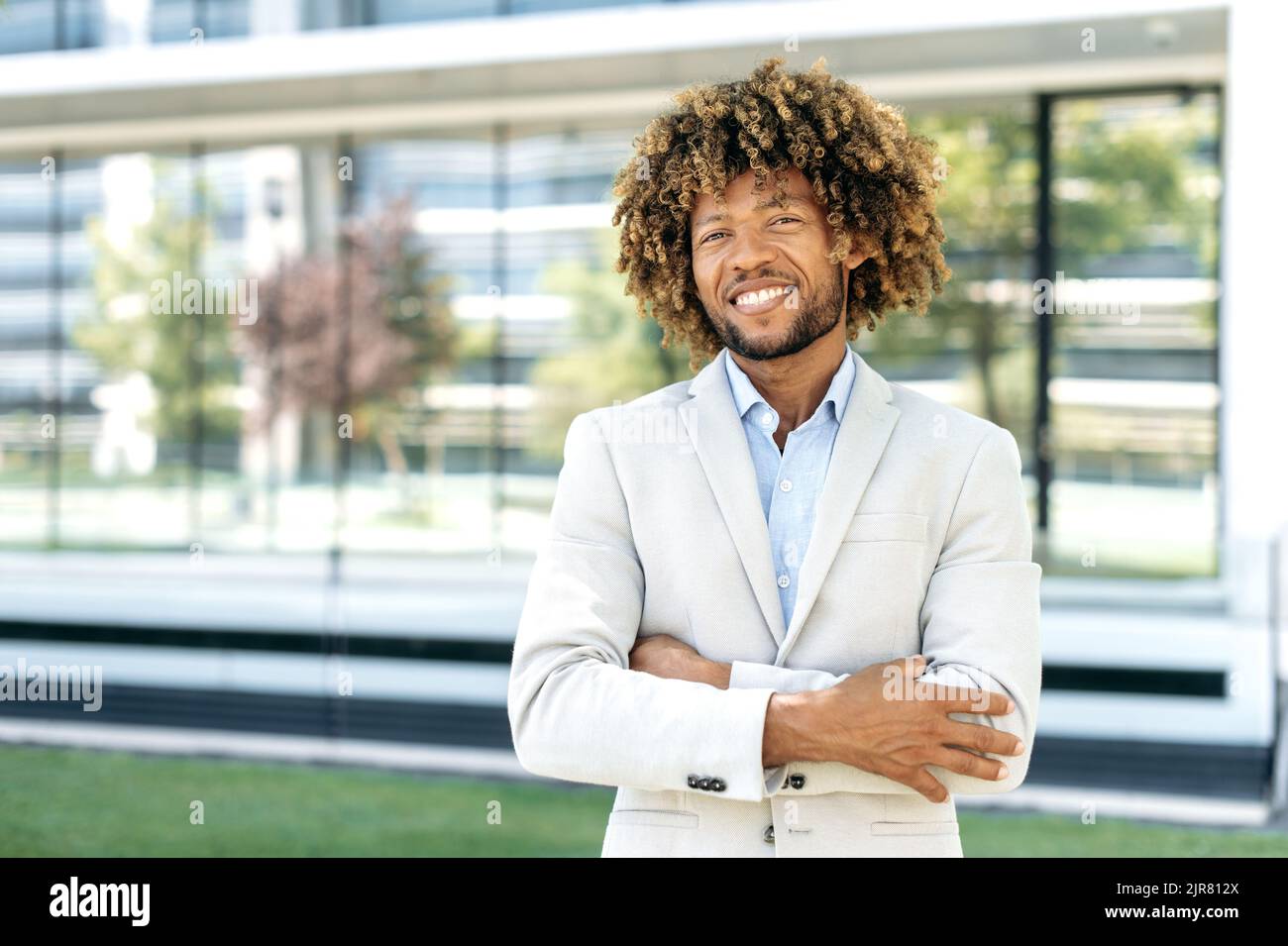 Elegante ritratto di un felice uomo brasiliano o ispanico con capelli ricci sicuro di successo, in piedi all'aperto con le braccia incrociate vicino al business center, guardando la macchina fotografica, sorridente amichevole Foto Stock