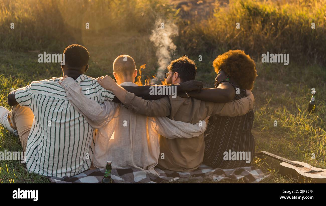 Vista posteriore di buoni vecchi amici multiculturali abbracciare mentre si siede su una coperta sull'erba, avendo un picnic di fine settimana nella natura vicino al fiume, parlando e bevendo la birra. Ora del tramonto. Foto di alta qualità Foto Stock
