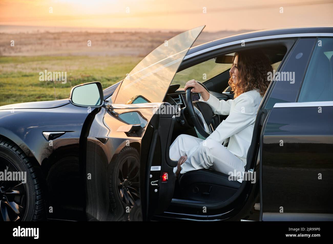 Vista laterale della donna allegra padrona di agriburaio seduto in auto nera e sorridente mentre si pensa alla proprietà della terra in ambiente naturale, donna Caucaisan felice in automobile godendo stile di vita Foto Stock