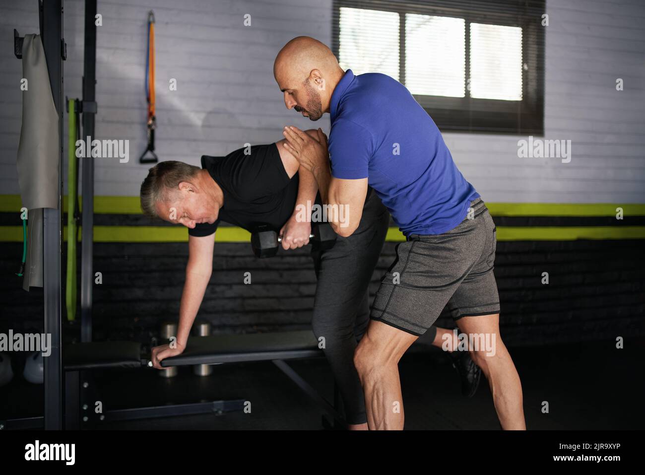Un personal trainer maschile assiste un anziano adulto che tiene la forma corretta nel fare file di braccio da banco. Addestramento di forza ed equilibrio per il rehemi Foto Stock