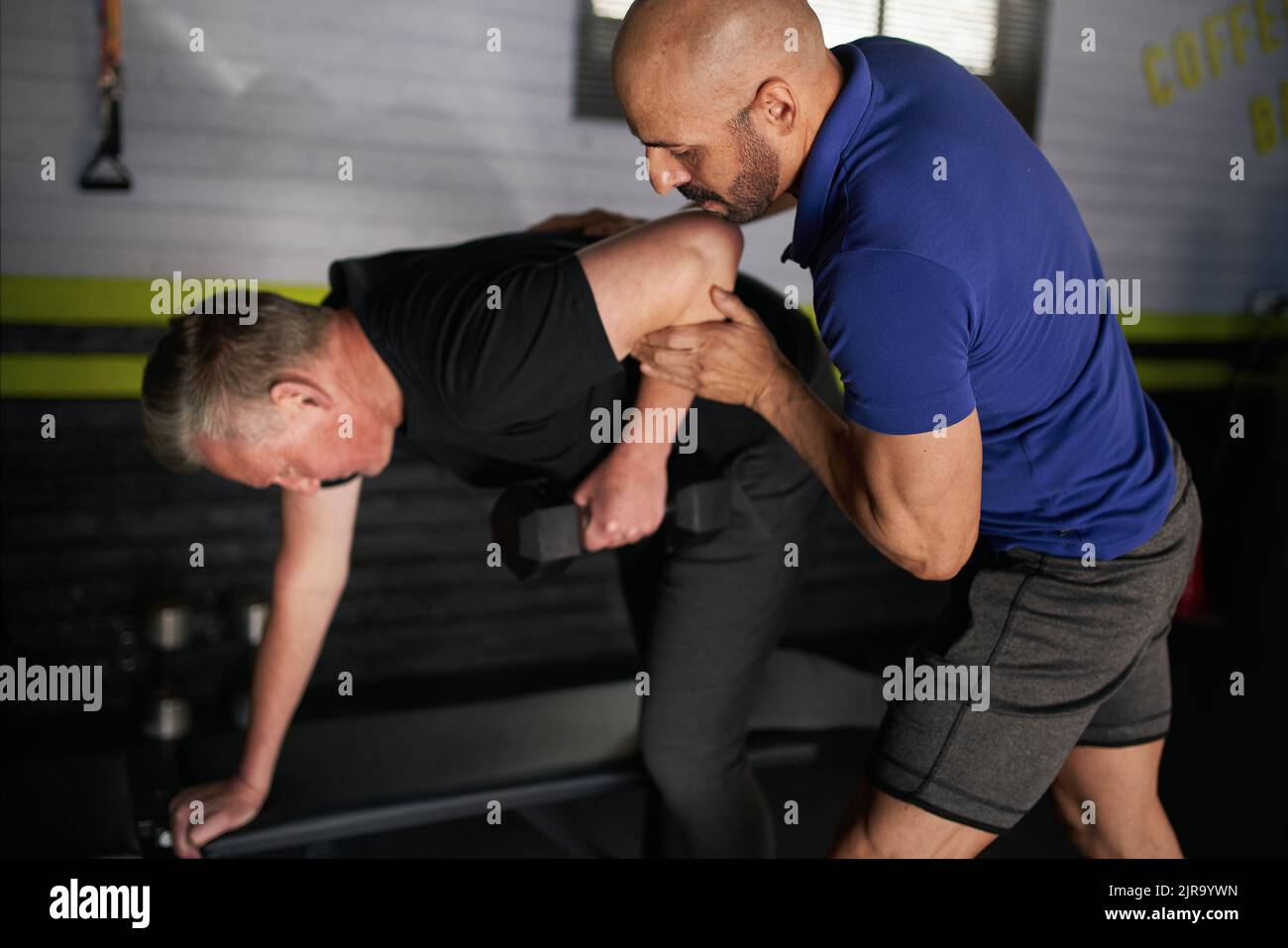 Primo piano di un personal trainer maschile che assiste un uomo adulto maturo nel mantenere la forma corretta nel fare file di braccio da banco. Allenamento per la forza e l'equilibrio per re Foto Stock
