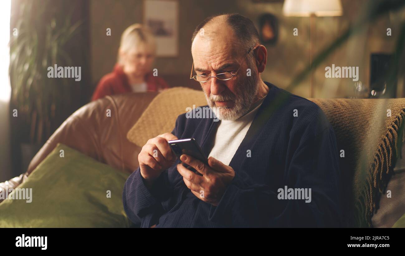 L'uomo anziano depresso sul divano togliendo gli occhiali mentre tiene il telefono con le notizie difettose e preoccupato Foto Stock