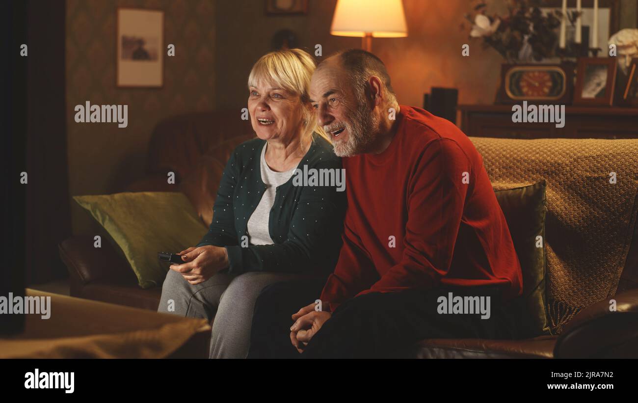 Allegra coppia anziana che ride e parla mentre si siede sul divano e guarda film divertenti in TV nel fine settimana sera a casa Foto Stock