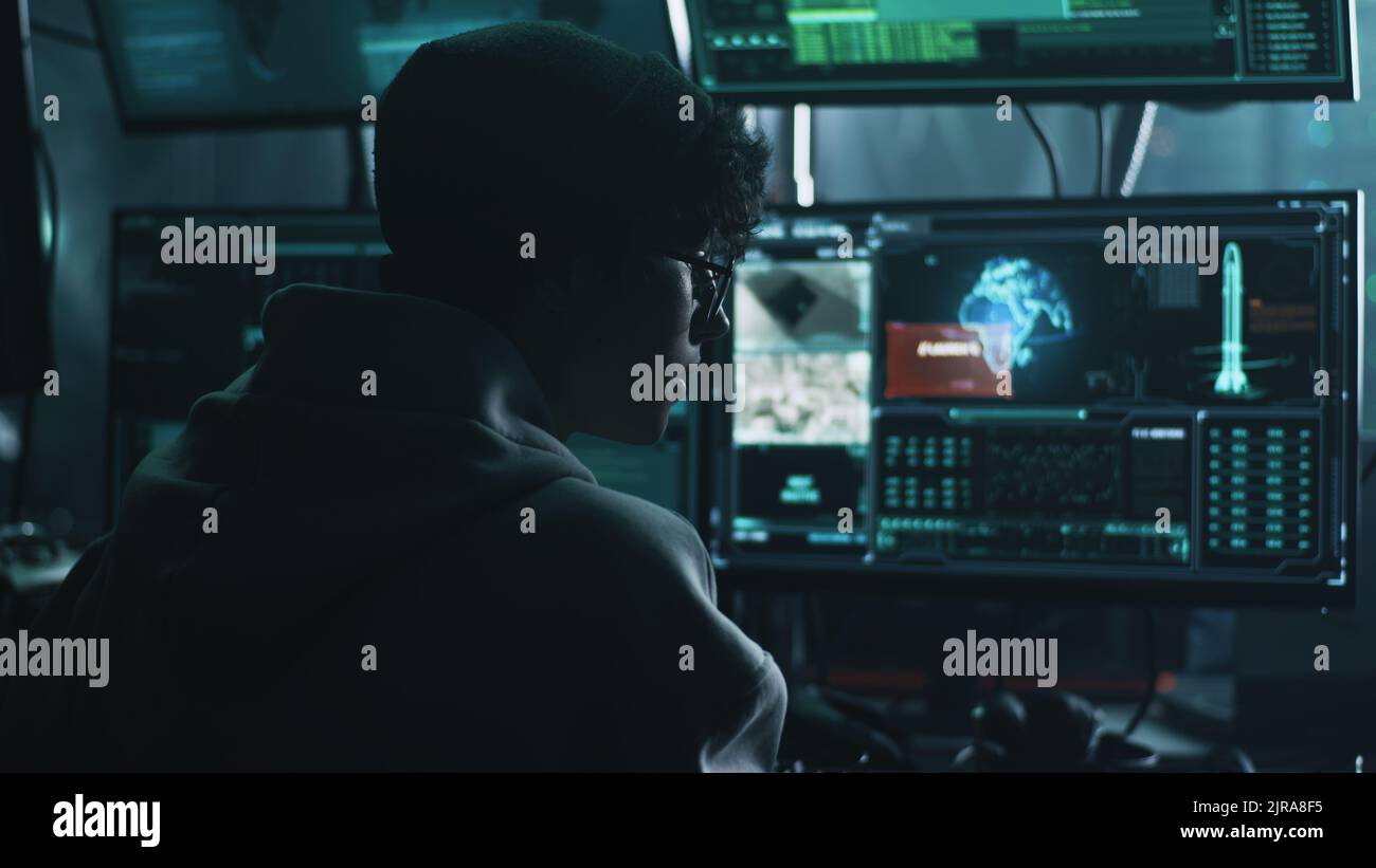 Un giovane hacker maschio in cappello e occhiali usando computer per hack nucleare testata e iniziare la guerra mentre si siede al tavolo in stanza buia Foto Stock