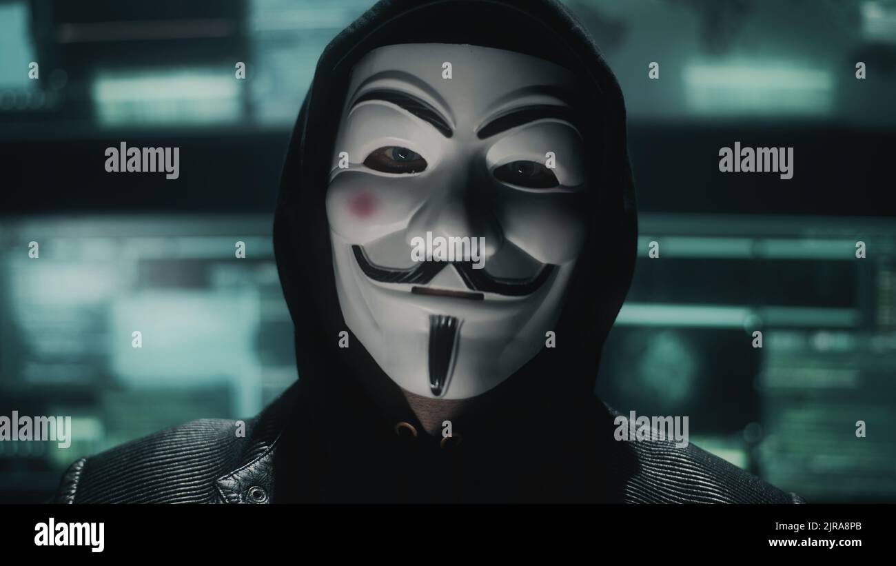 Hacker web maschile nel cappuccio e maschera anonima guardando la telecamera durante l'hacking di dati e server privato in un nascondiglio oscurato Foto Stock