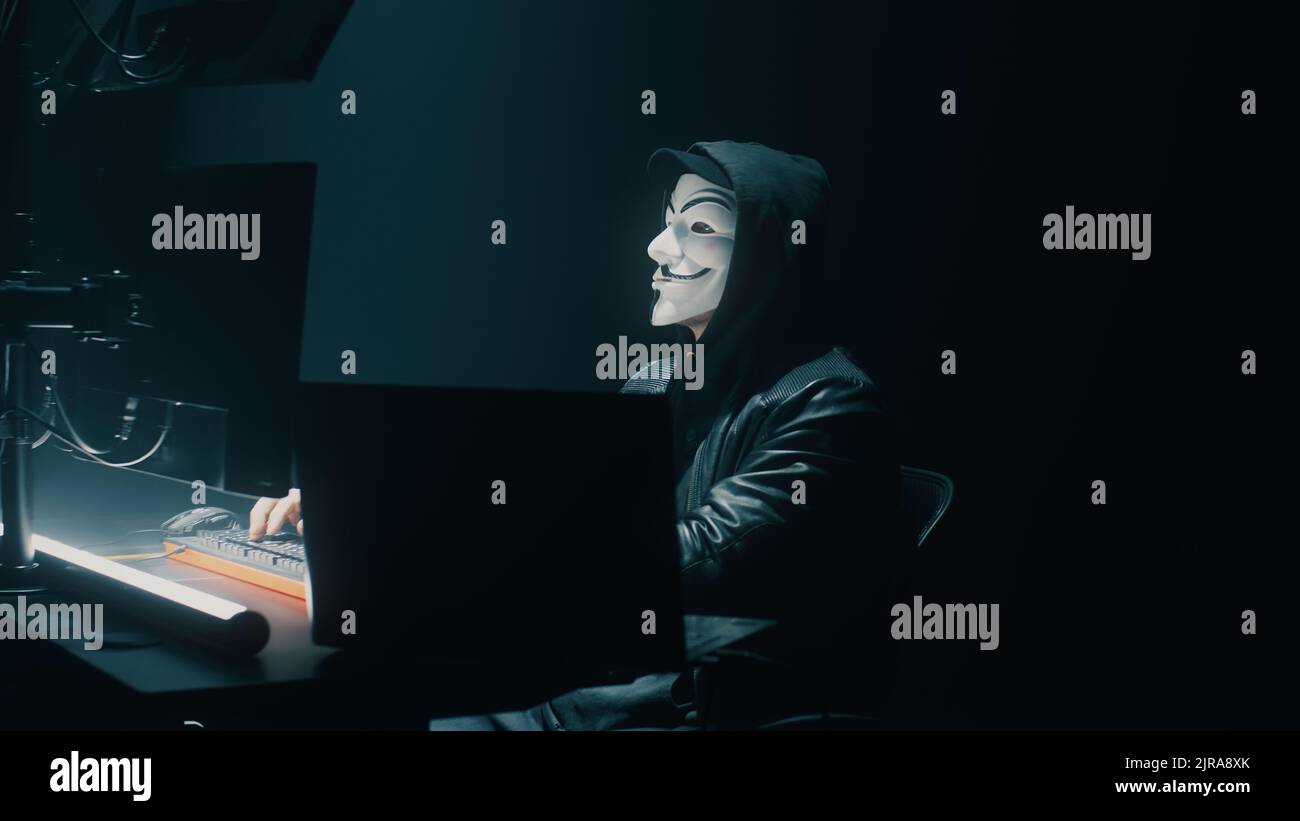 Frode irriconoscibile nella maschera usando il computer per rubare i dati nella stanza buia di notte Foto Stock