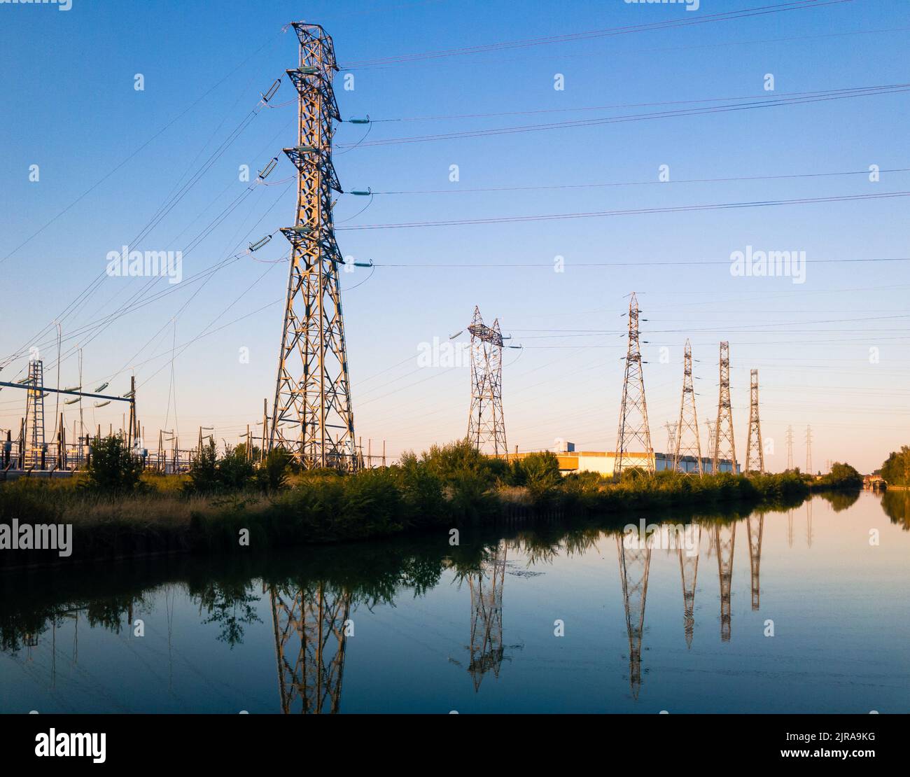 Una fila di torri metalliche di trasmissione di varie forme che si riflettono nell'acqua ferma di un canale contro il cielo blu al crepuscolo. Foto Stock