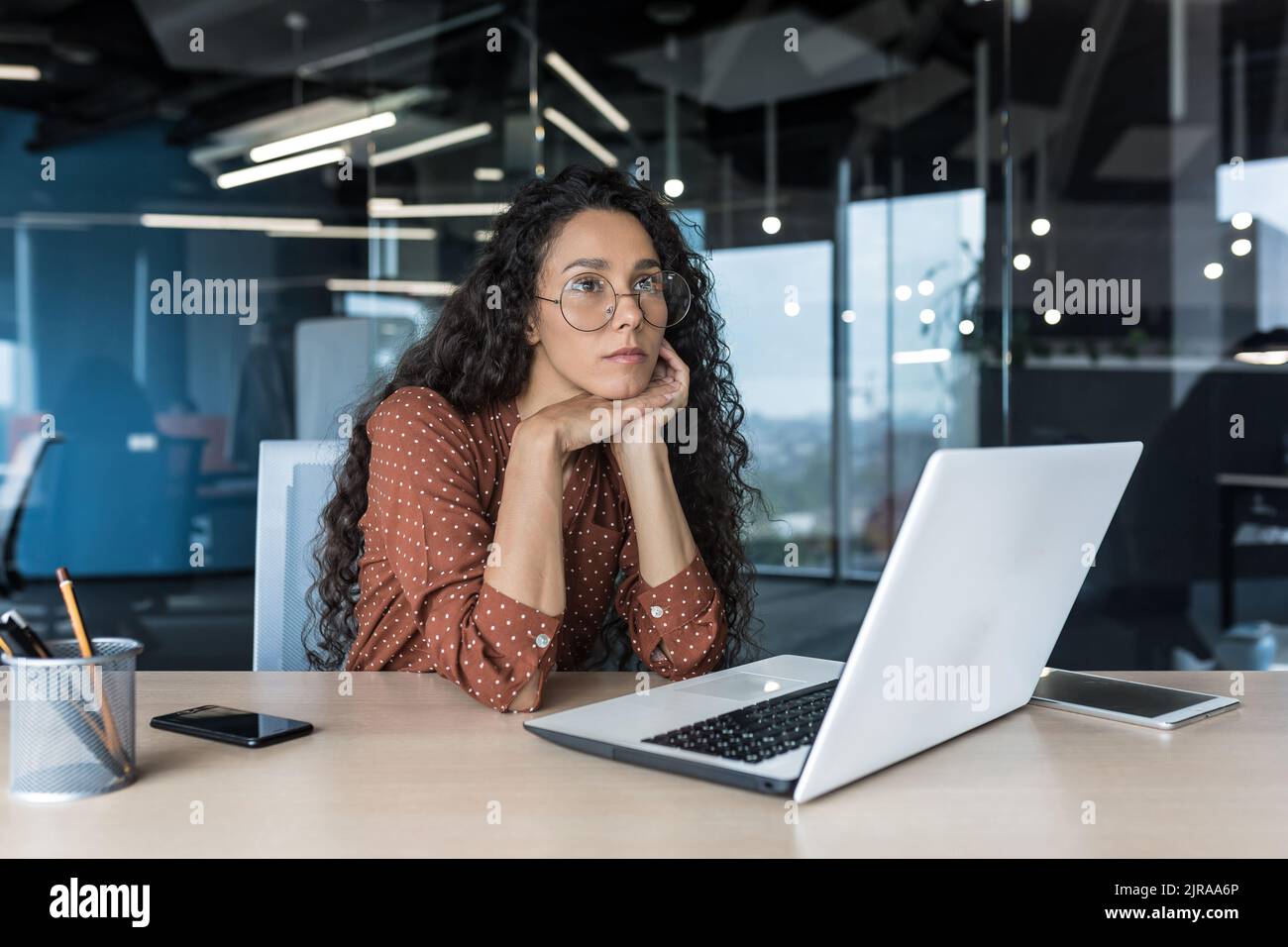 Giovane donna ispanica bella con capelli ricci e occhiali, che lavora in ufficio moderno utilizzando il computer portatile, il pensiero di lavoro femminile e triste, donna di affari sconvolto Foto Stock