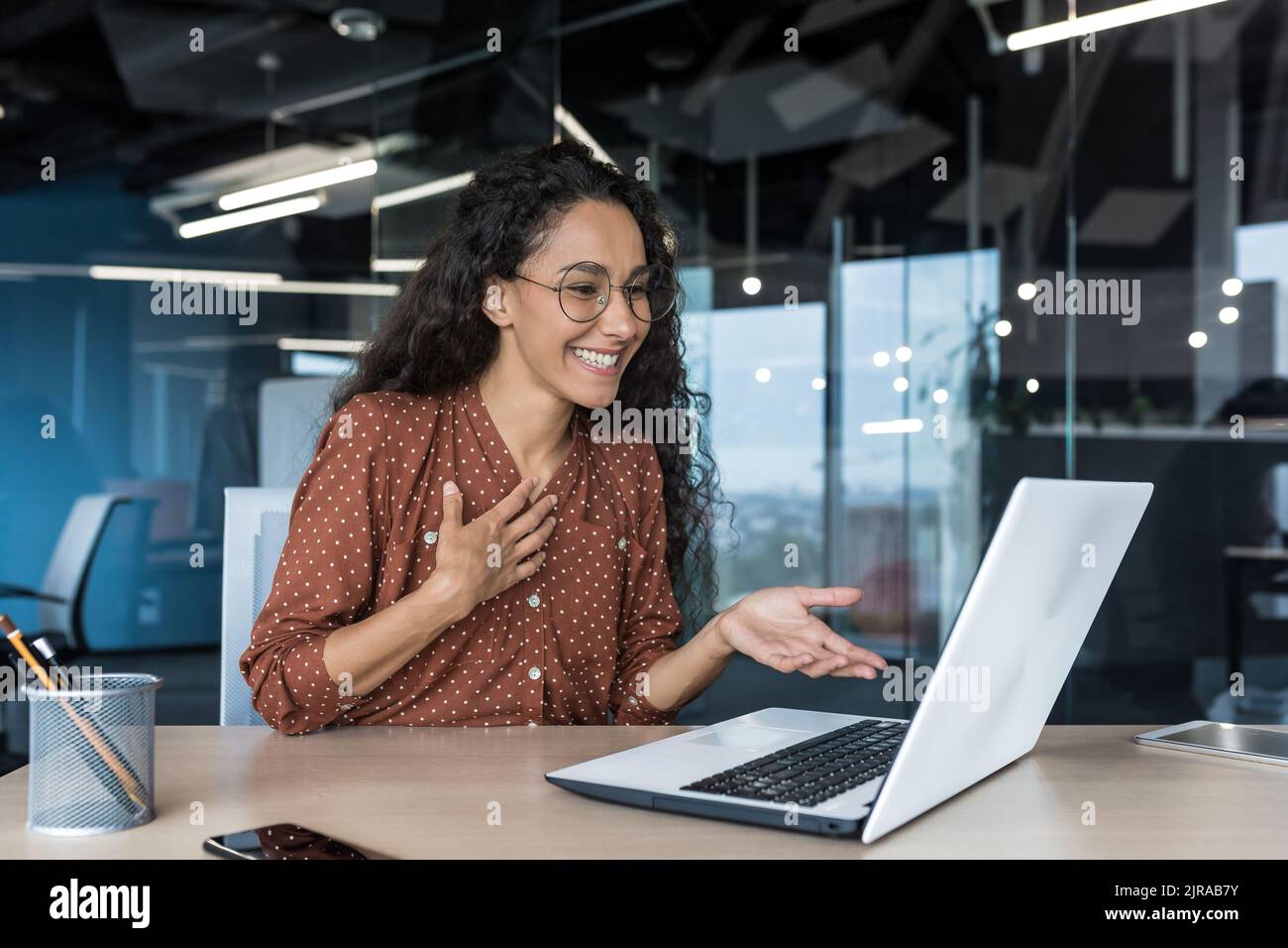 Donna d'affari ispanica felice che lavora in un ufficio moderno utilizzando un computer portatile per videochiamare e riunioni online con colleghi dipendenti, donna sorridente e divertente dando una presentazione Foto Stock