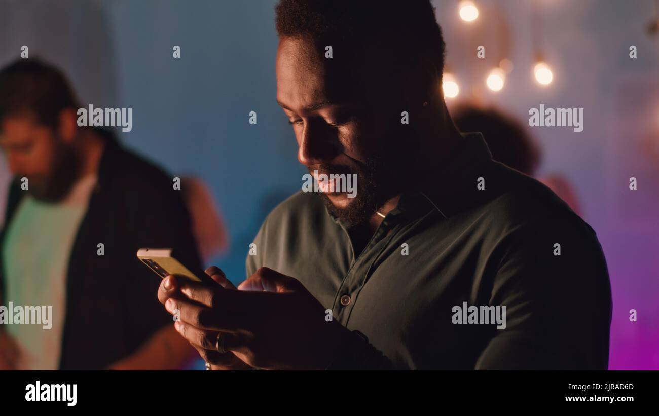 Uomo afro-americano ottimista con alcol coctail lettura e l'invio di messaggi di testo sul cellulare durante la festa a casa in camera piena di fumo Foto Stock