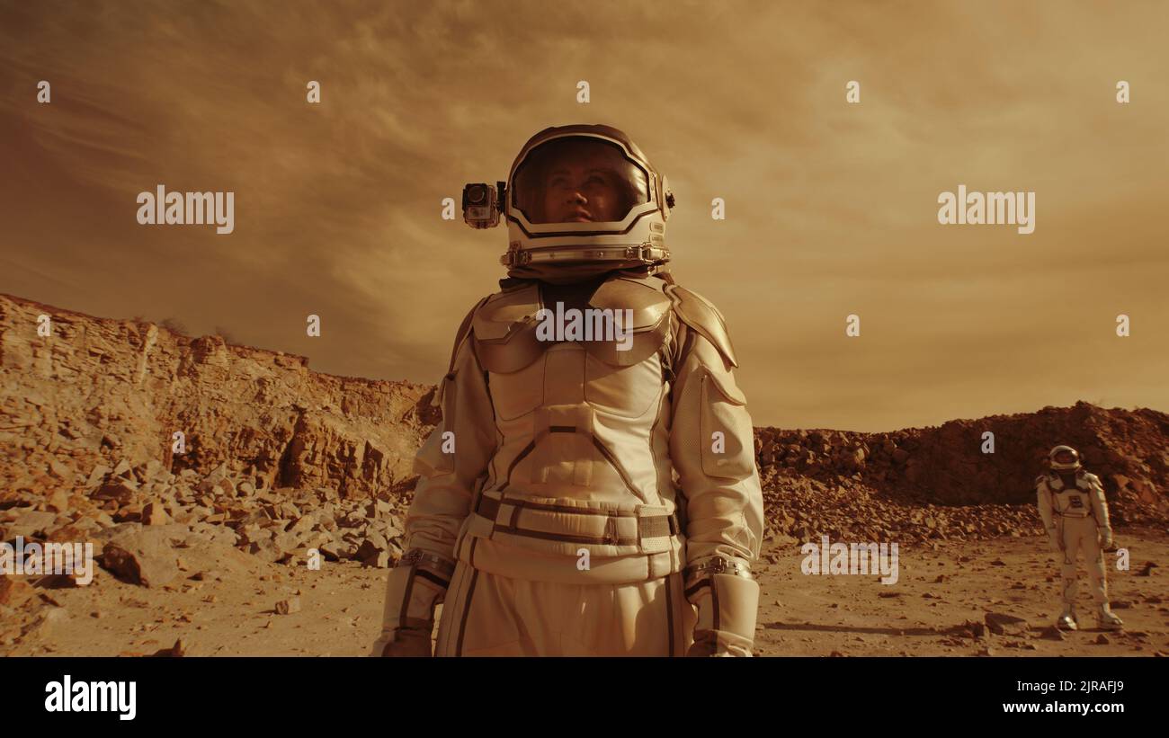 Panoramica della donna in una tuta spaziale guardando lontano e osservando il terreno roccioso mentre si è in piedi vicino a un collega durante la colonizzazione di Marte Foto Stock