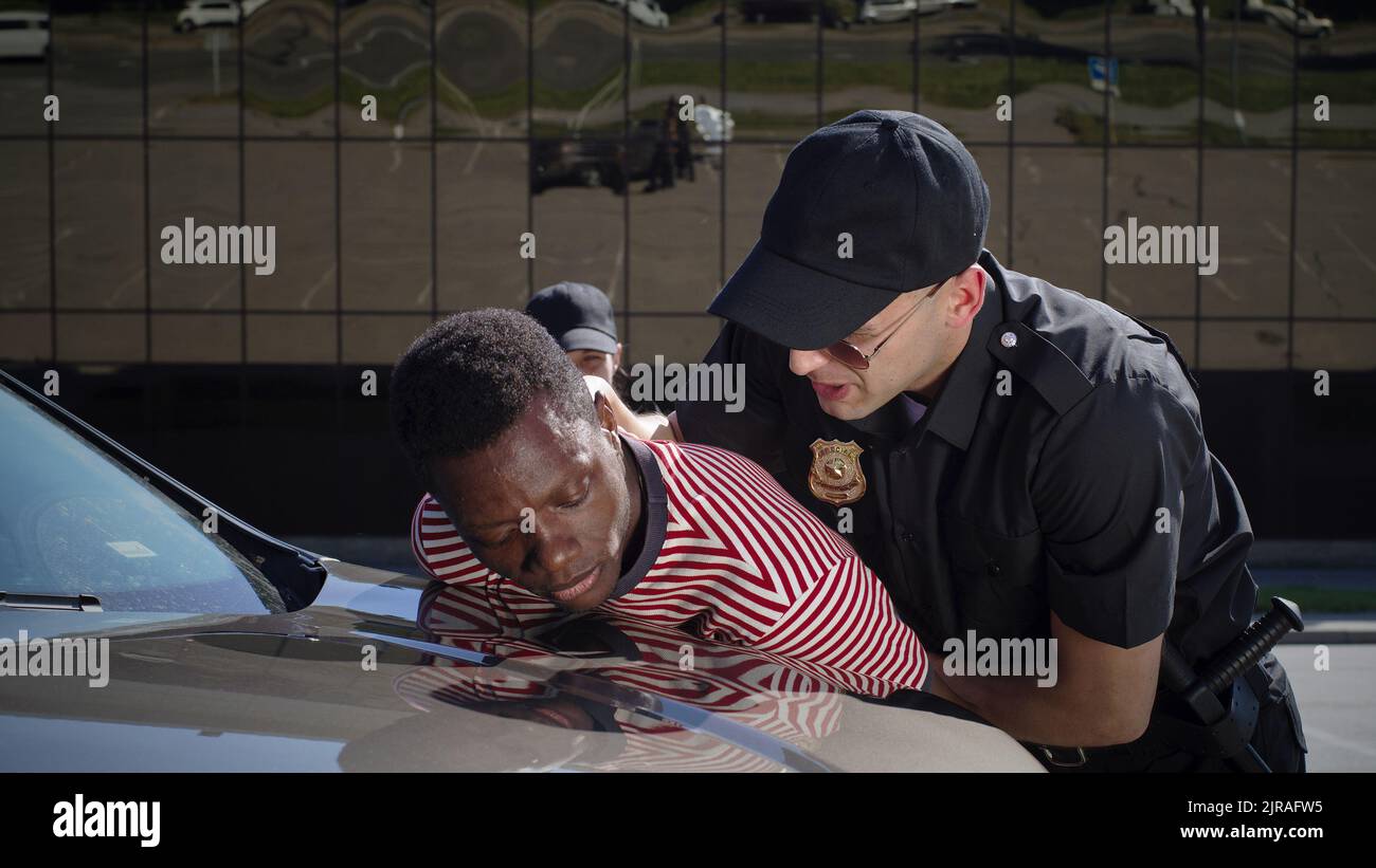 Uomo e donna in uniforme di polizia puntando pistola e cercando resistente afro-americano ragazzo vicino auto durante l'arresto Foto Stock