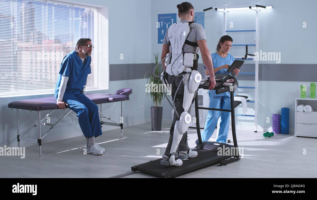 Uomo e donna in uniforme medica che controlla tapis roulant e discutere i risultati mentre il paziente maschile in esoscheletro imparare a camminare durante la sessione di riabilitazione in ospedale futuristico Foto Stock