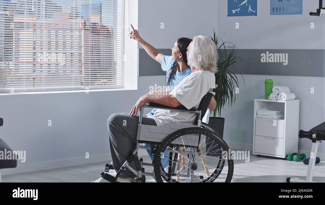 Donna adulta medico sorridente e indicando la città dietro la finestra mentre si parla con l'anziano con disabilità durante il lavoro in ospedale riabilitazione Foto Stock