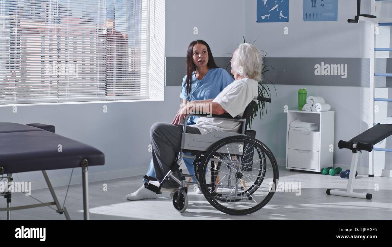 Donna adulta medico sorridente e indicando la città dietro la finestra mentre si parla con l'anziano con disabilità durante il lavoro in ospedale riabilitazione Foto Stock