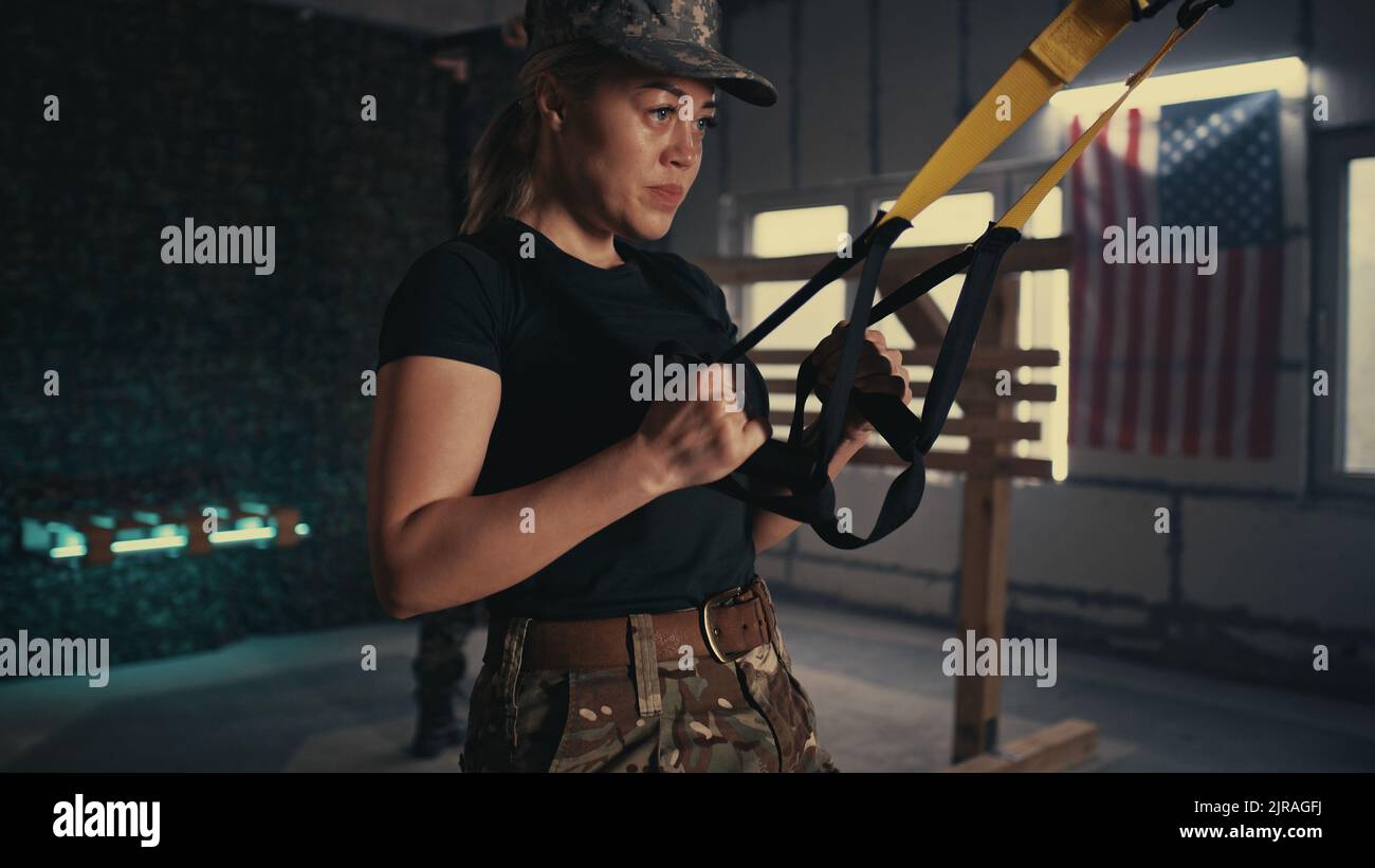 Vista della donna stanca in pantaloni camouflage facendo TRX affondi vicino squadmate durante l'allenamento in palestra scura della base militare degli Stati Uniti Foto Stock