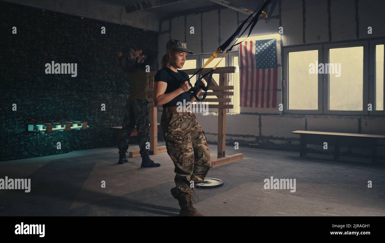 Vista della donna stanca in pantaloni camouflage facendo TRX affondi vicino squadmate durante l'allenamento in palestra scura della base militare degli Stati Uniti Foto Stock