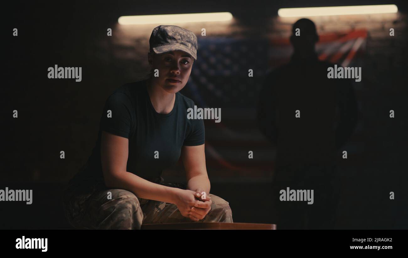 Donna in mimetizzazione seduta su panca e guardando la telecamera contro la bandiera degli Stati Uniti e la silhouette del comandante su base militare Foto Stock