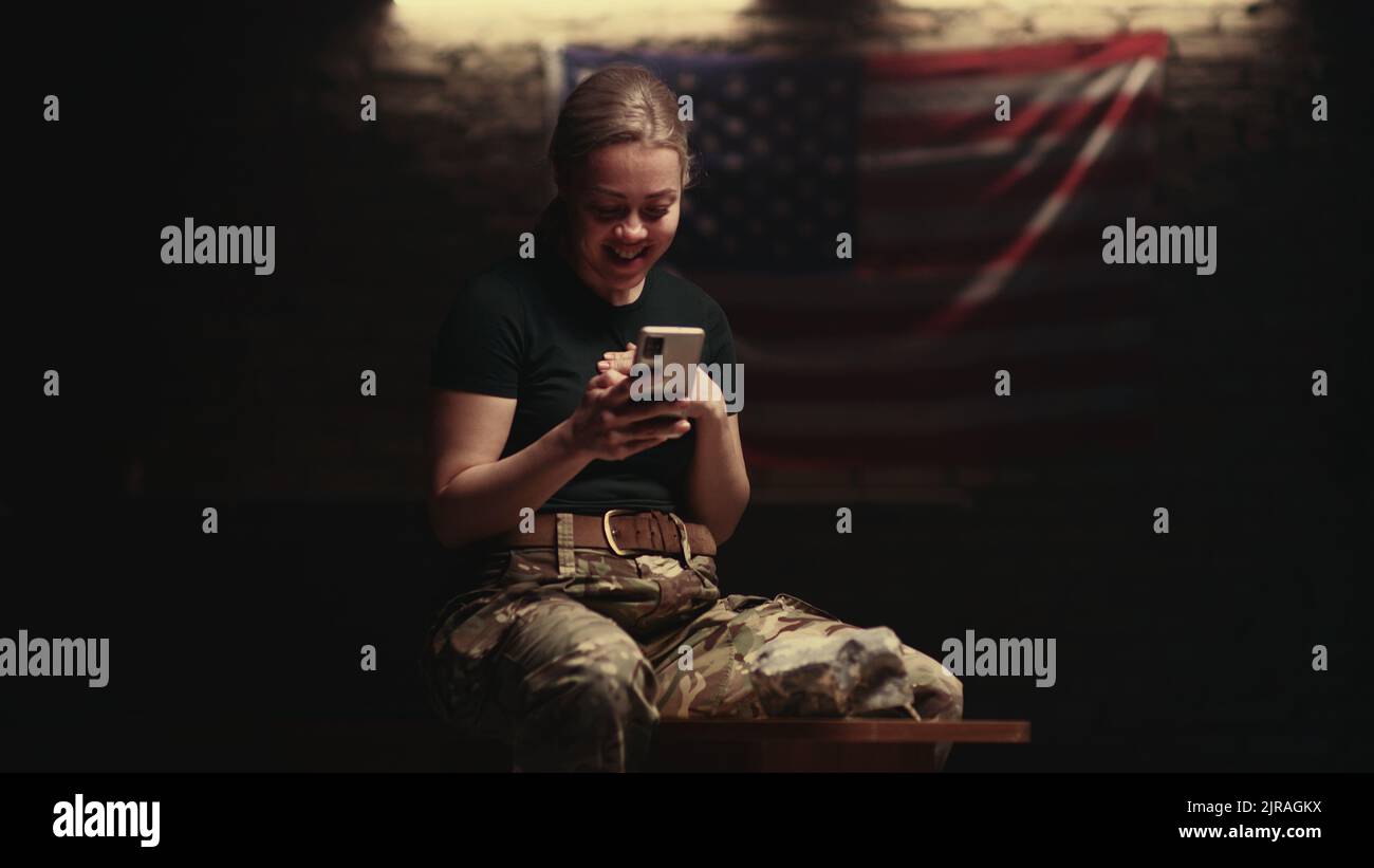 Donna ottimista nel mimetismo sorridendo e parlando con un amico online mentre si siede sulla panchina nella stanza oscura della base militare degli Stati Uniti Foto Stock