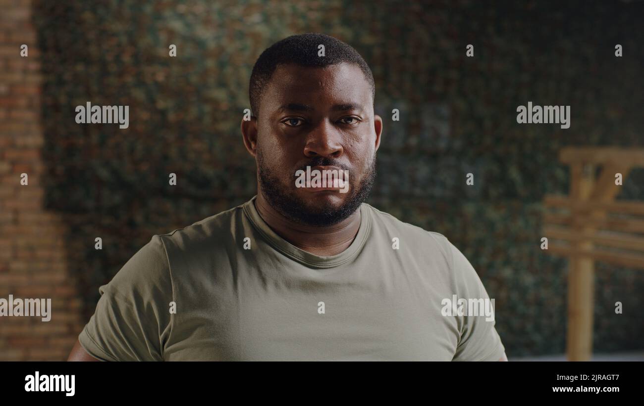 Uomo militare bearded guardando la macchina fotografica con faccia seria contro il muro di mattoni con la bandiera degli Stati Uniti sulla base militare Foto Stock