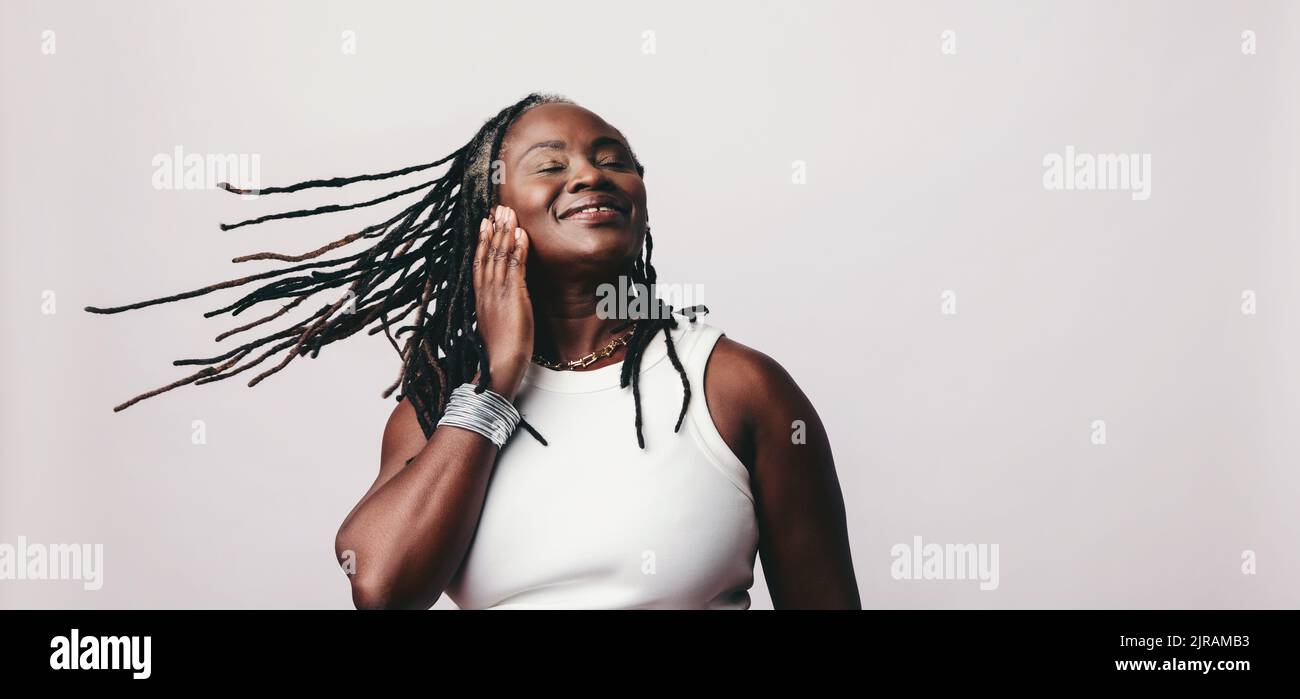 Donna felice che frusta i suoi Dreadlock mentre ascolta musica vibrante sugli auricolari. Donna matura allegra che ama la sua playlist preferita mentre è in piedi Foto Stock