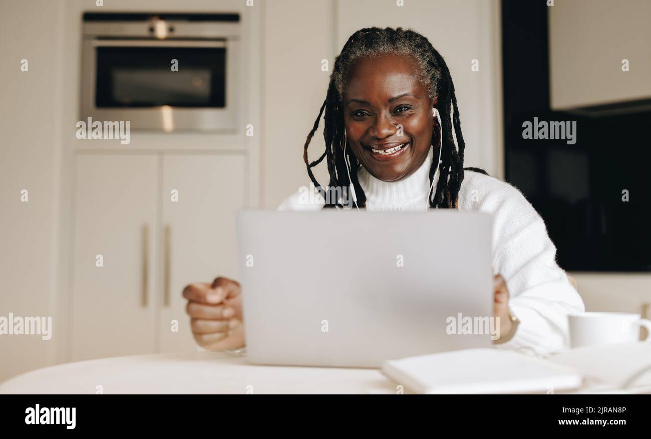 Felice donna d'affari senior sorridente alla fotocamera mentre lavora su un notebook nel suo ufficio di casa. Donna d'affari matura che ascolta musica mentre lavora f Foto Stock