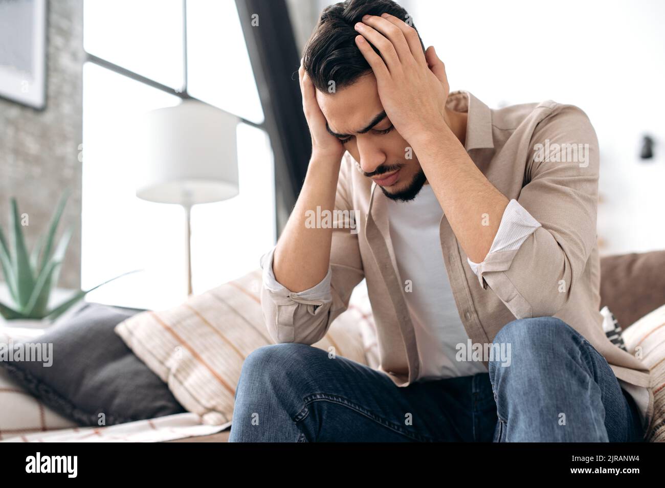 Frustrato uomo indiano o arabo triste, sedersi a casa sul divano nel soggiorno con gli occhi chiusi, tenendo le mani sulla testa, soffrendo di mal di testa, sperimentando problemi personali. Cefalea, emicrania Foto Stock