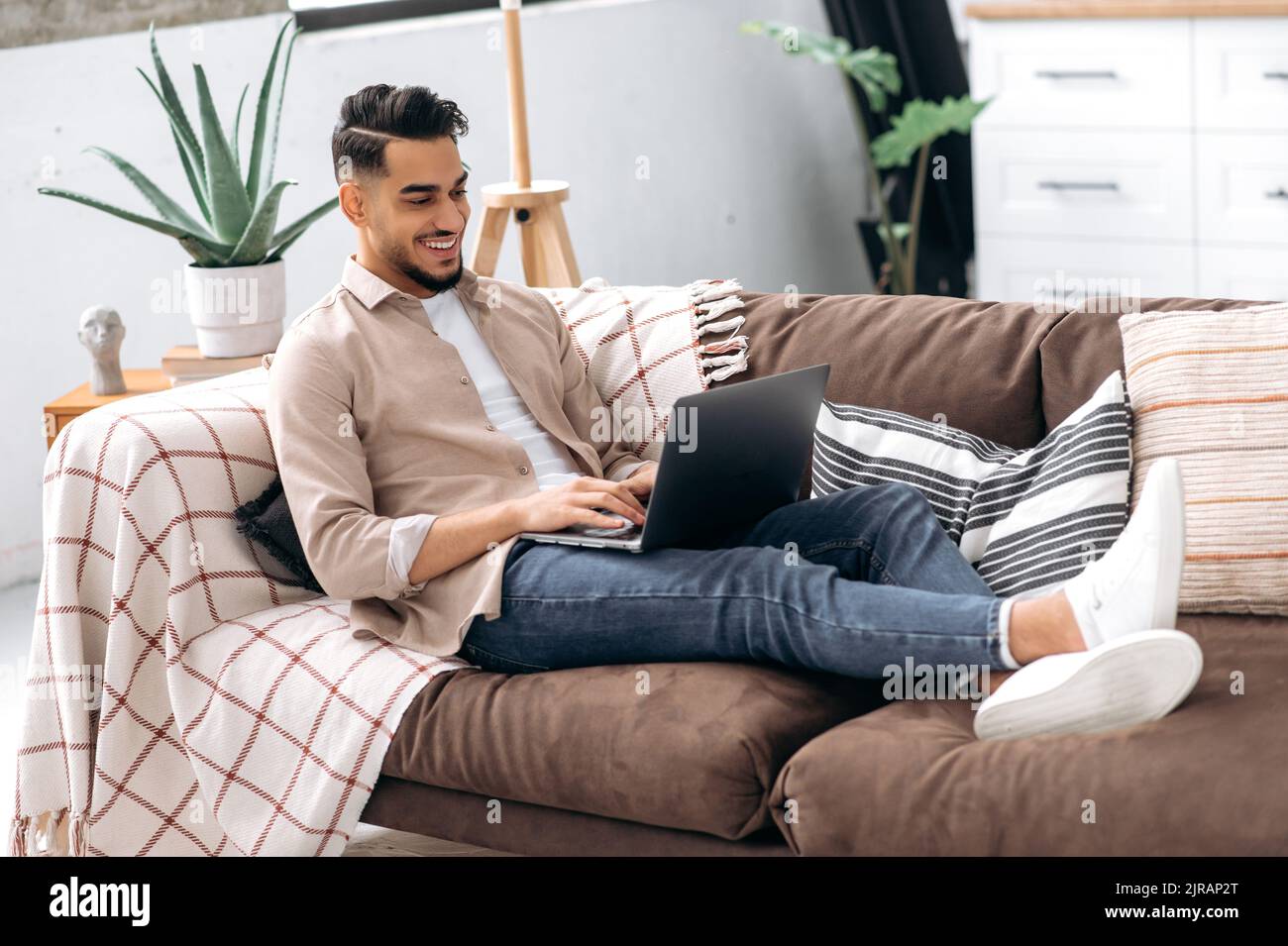 Giovane arabo o indiano positivo in abiti casual elegante, freelancer, web designer, seduto sul divano in un salotto, utilizza il laptop per lavorare su un progetto, elearning, navigare in internet, sorridere Foto Stock
