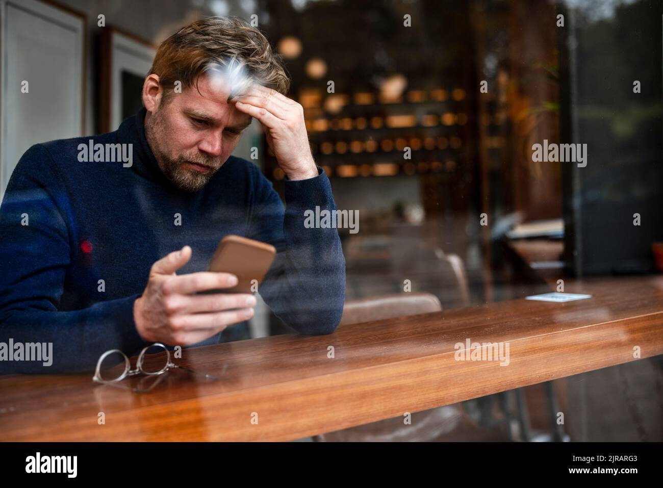 Un uomo d'affari preoccupato che usa lo smartphone in un bar visto attraverso una finestra di vetro Foto Stock