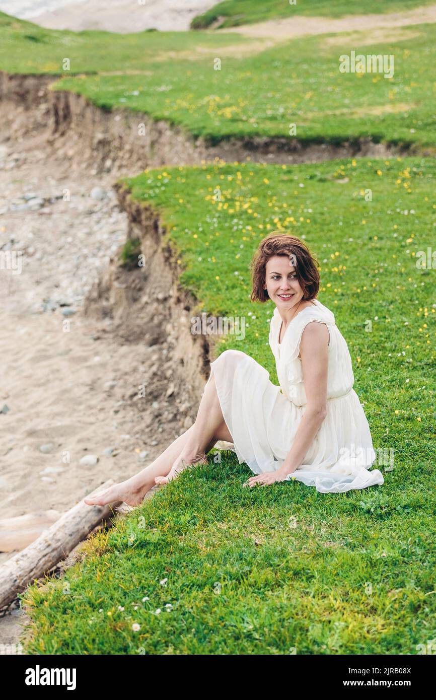 Donna sorridente che indossa un abito bianco seduto sull'erba Foto Stock
