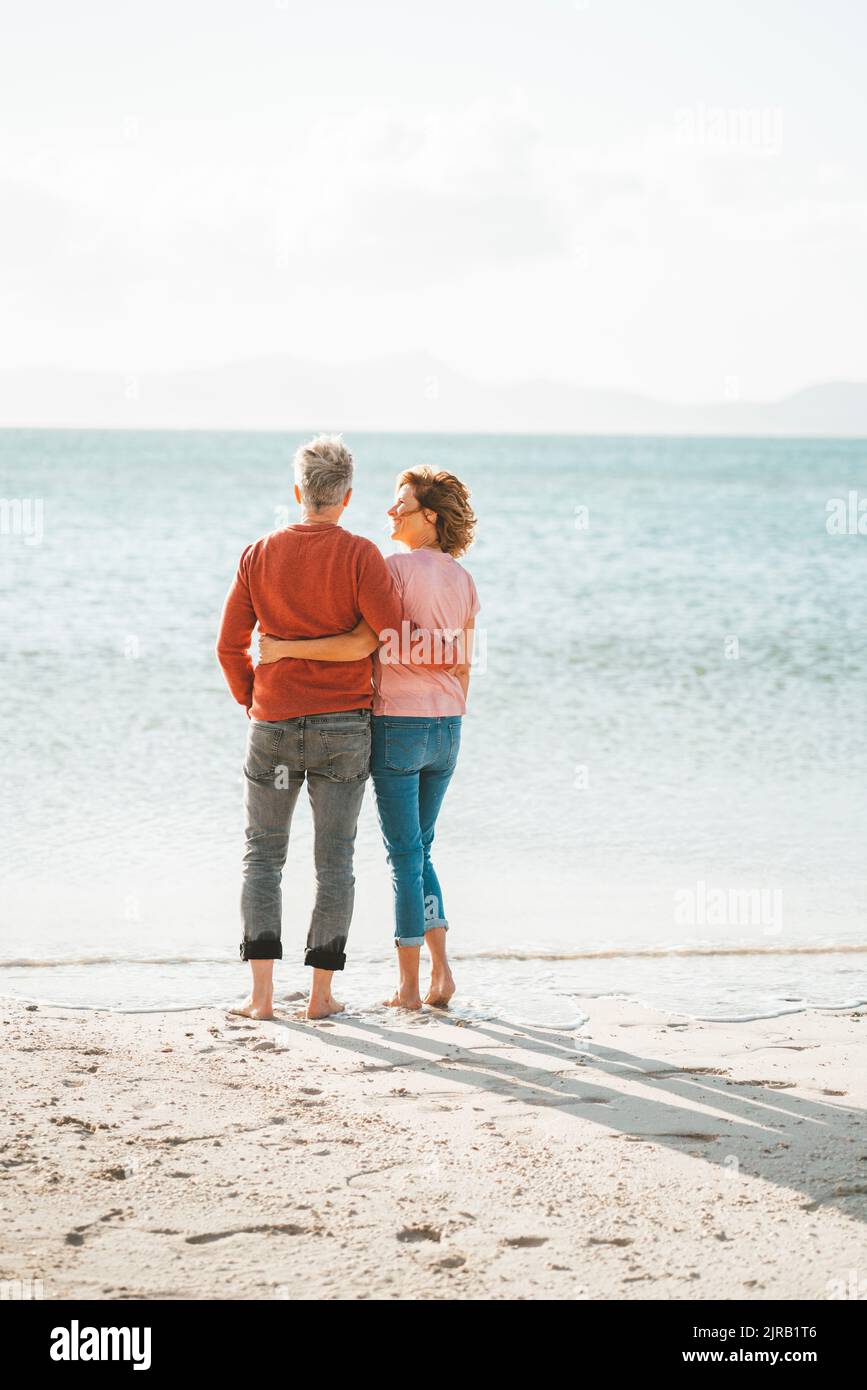 Coppia matura con le braccia intorno l'un l'altro in piedi sulla riva in spiaggia Foto Stock