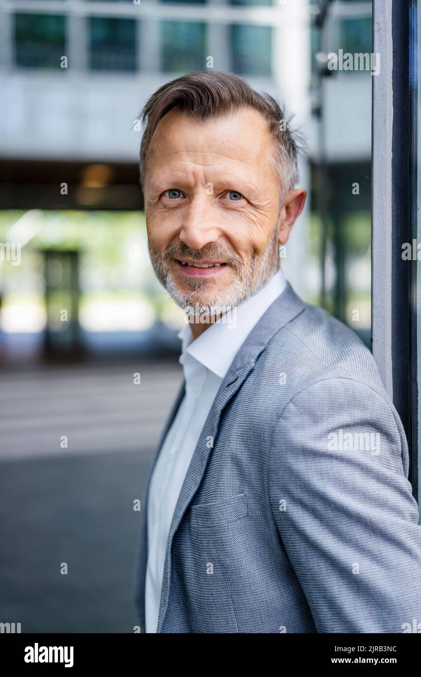 Uomo d'affari maturo sorridente in blazer grigio appoggiato al muro Foto Stock