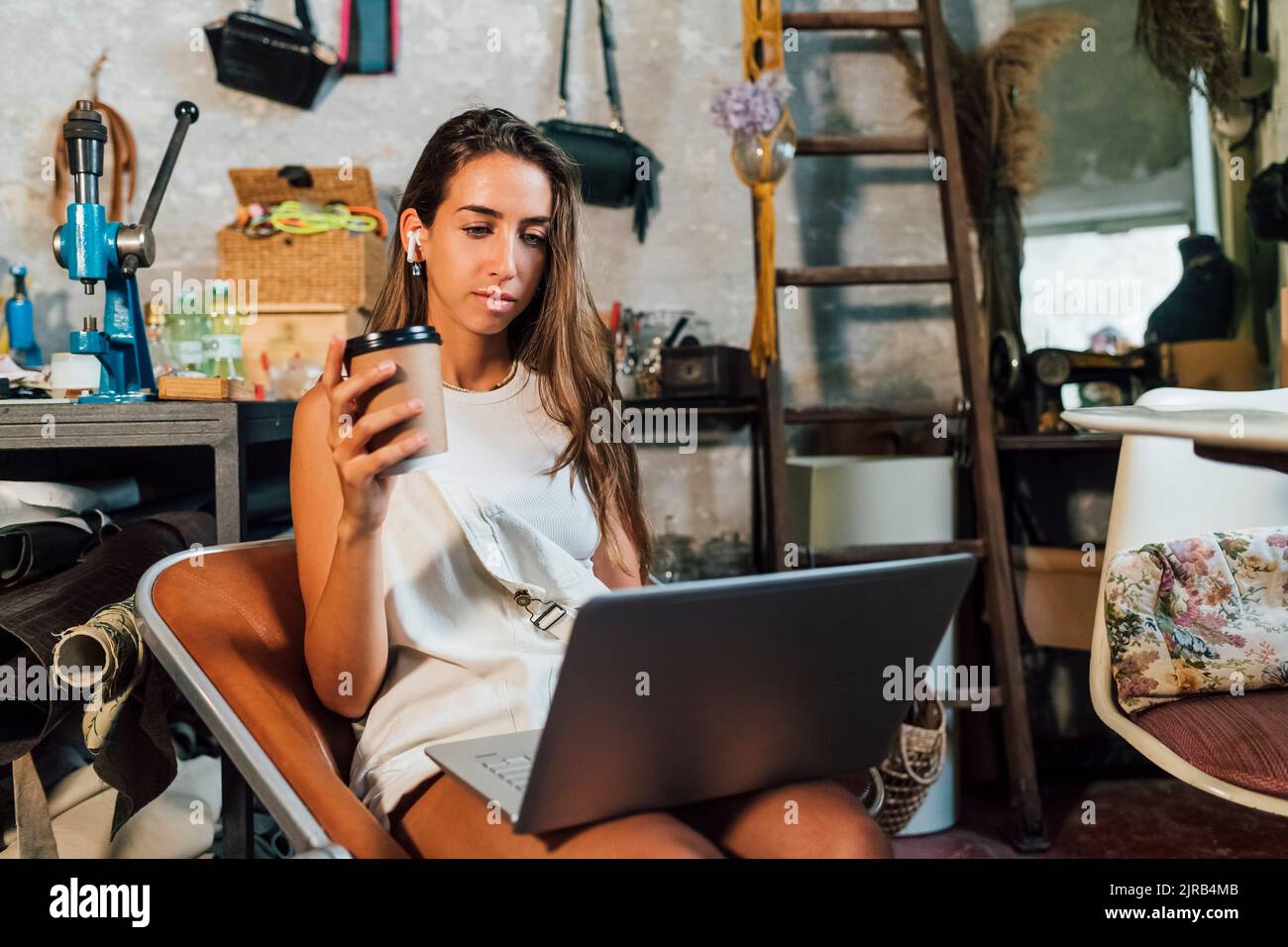 Stilista che tiene una tazza da caffè usa e getta utilizzando un computer portatile in officina Foto Stock
