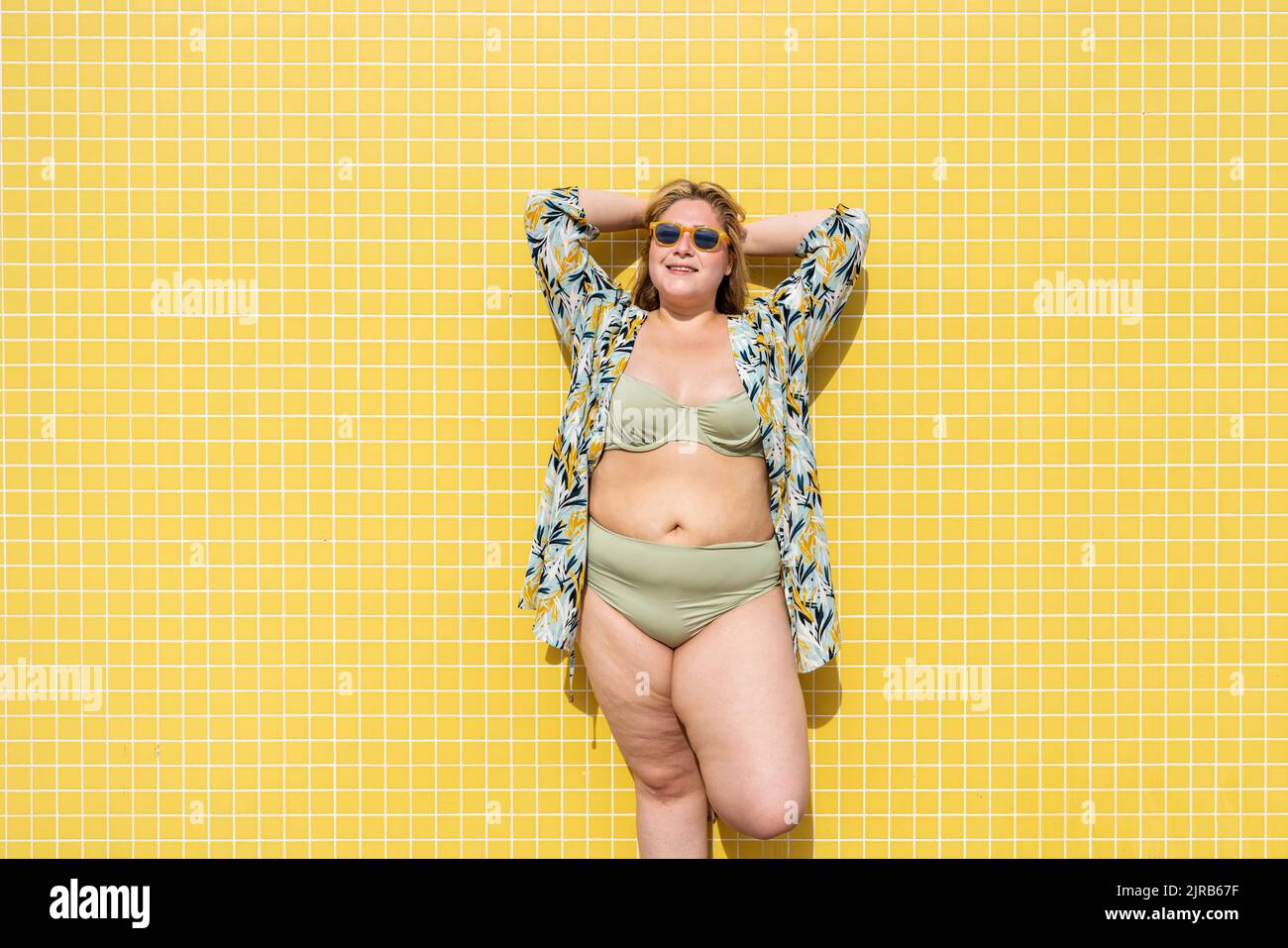 Donna sorridente più grande con le mani dietro la testa di fronte al muro giallo Foto Stock