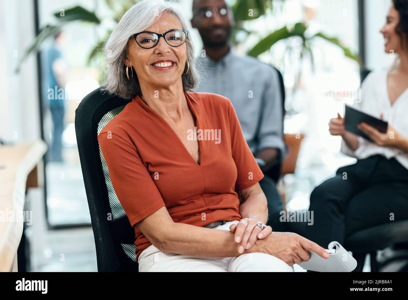 Donna d'affari anziana sorridente seduta sulla sedia sul posto di lavoro Foto Stock