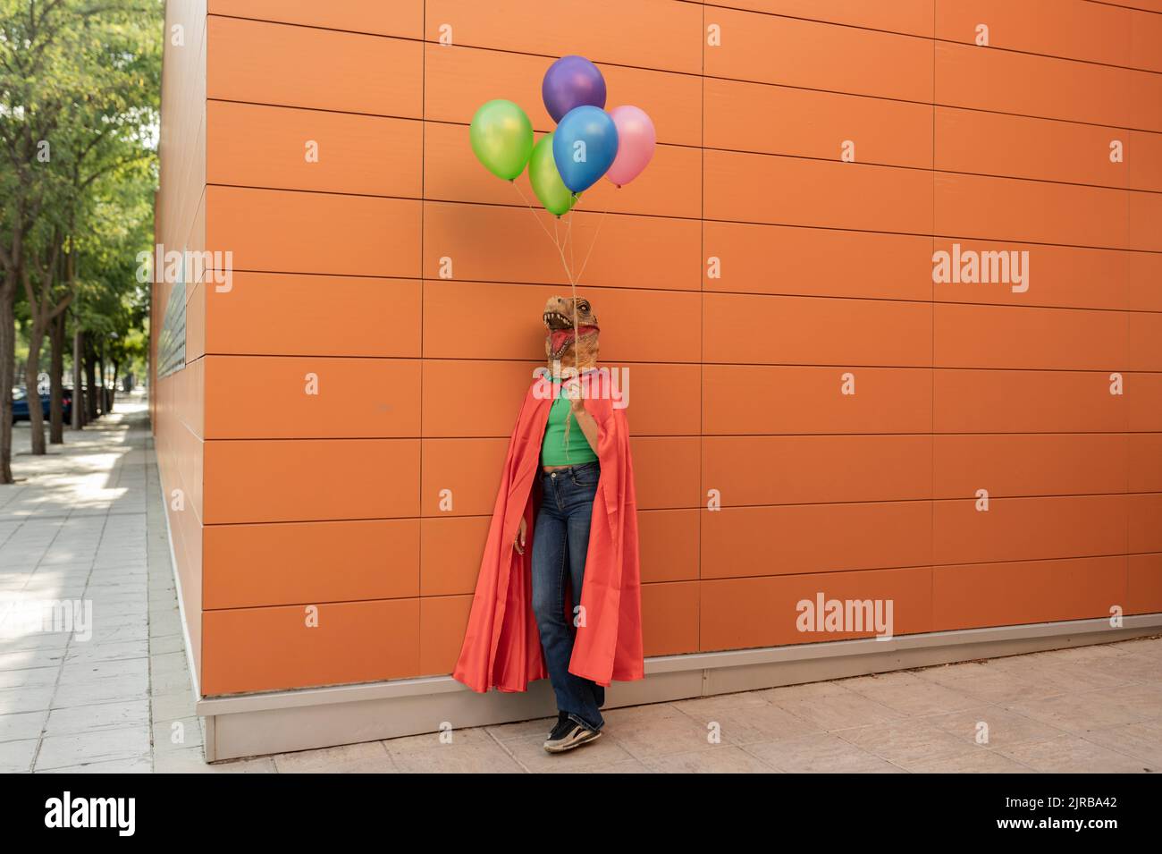 Giovane donna che indossa una maschera di coccodrillo e un mantello in piedi con palloncini multicolore davanti al muro Foto Stock