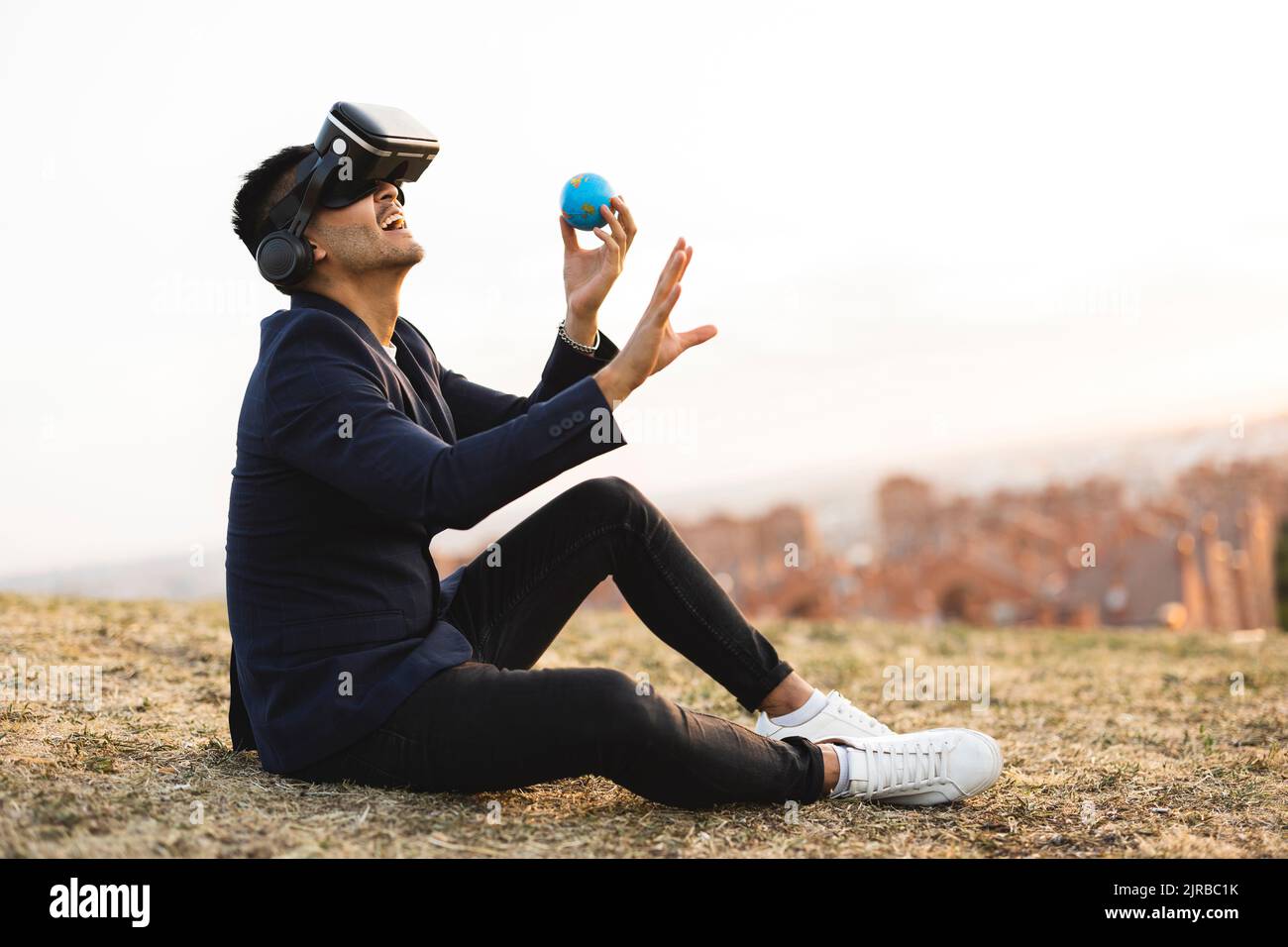 Giovane uomo d'affari con simulatore di realtà virtuale che tiene il globo seduto in collina Foto Stock