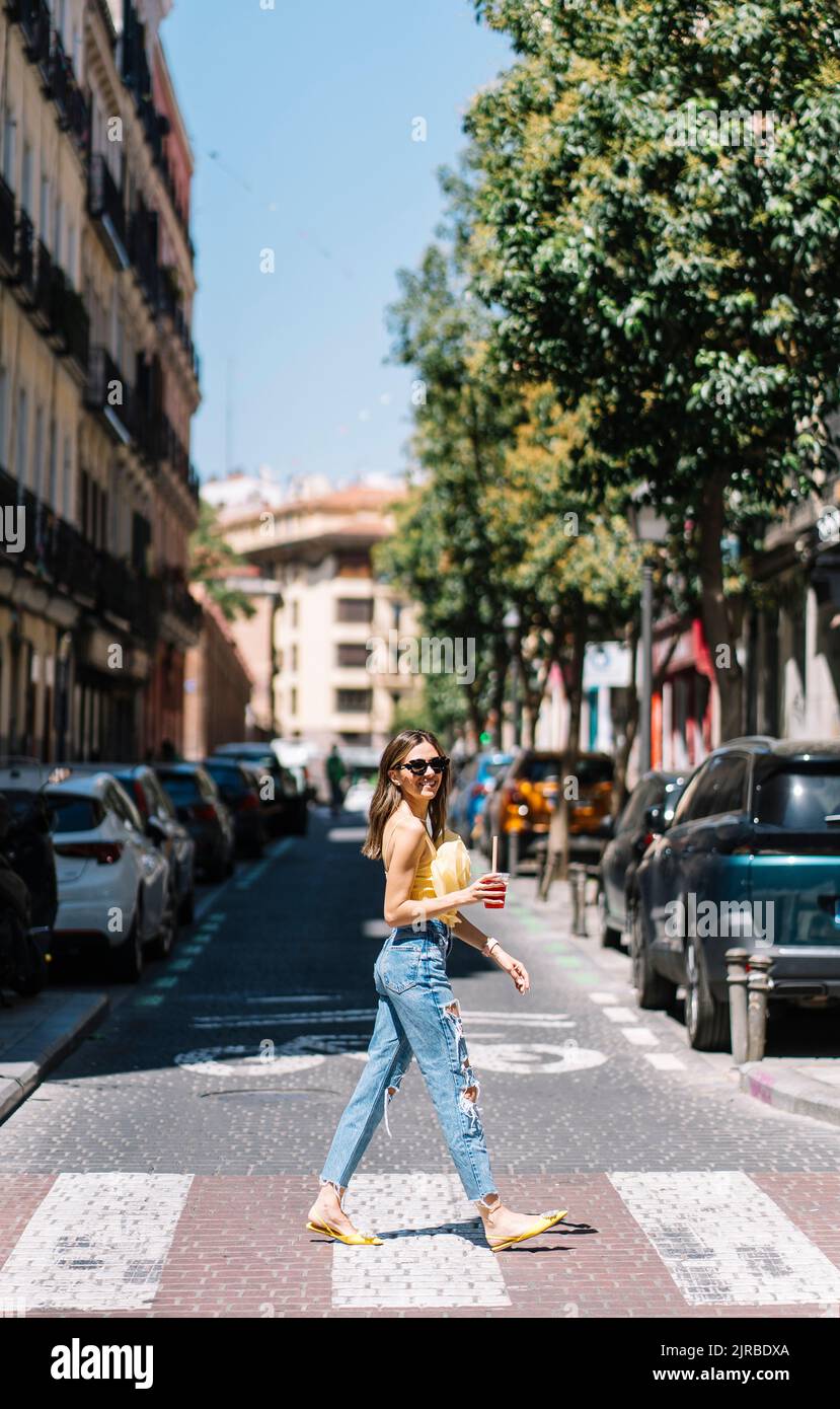 Giovane donna alla moda con drink in mano attraversando strada Foto Stock