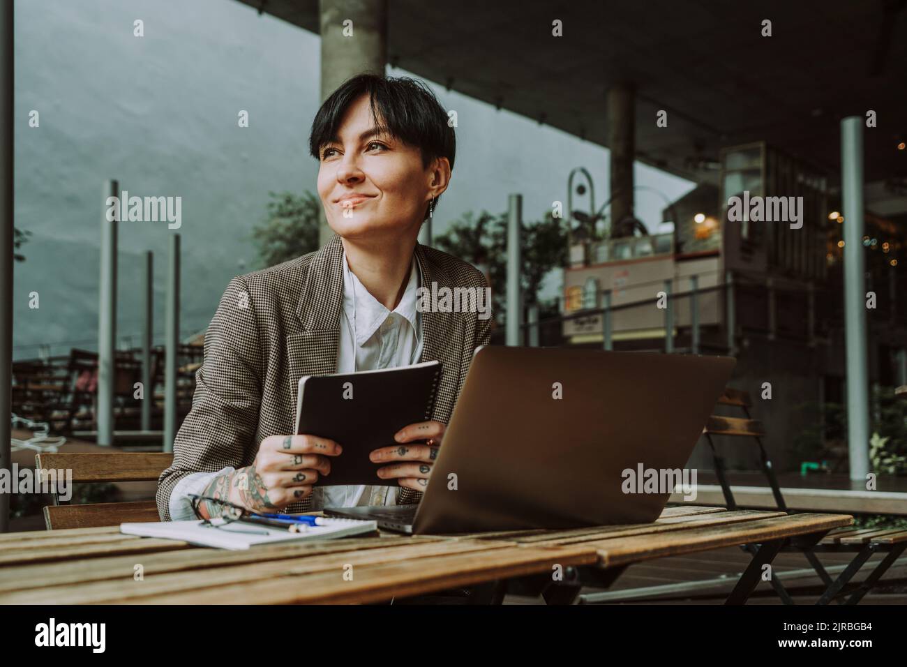 Donna d'affari sorridente con un computer portatile e un diario seduti al bar sul marciapiede Foto Stock