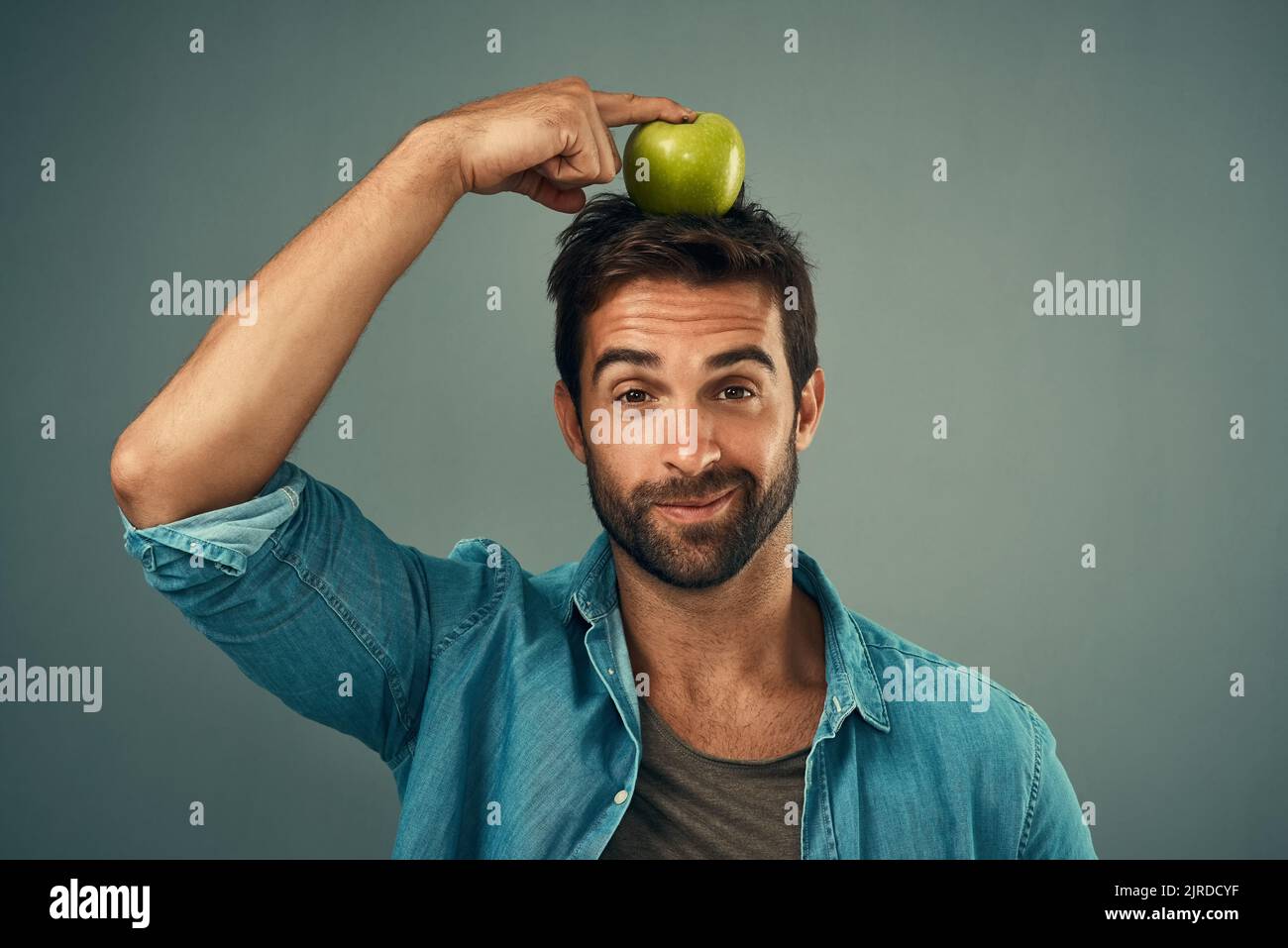 Il relativo circa trovare il giusto equilibrio. Studio ritratto di un bel giovane che posa con una mela sulla testa su uno sfondo grigio. Foto Stock