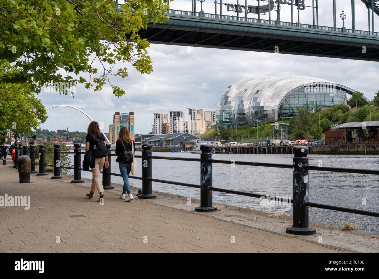 Due persone che camminano sul Quayside, Newcastle upon Tyne, Regno Unito, sotto il Tyne Bridge, con una vista dei luoghi di interesse di Gateshead, tra cui la Glasshouse Foto Stock