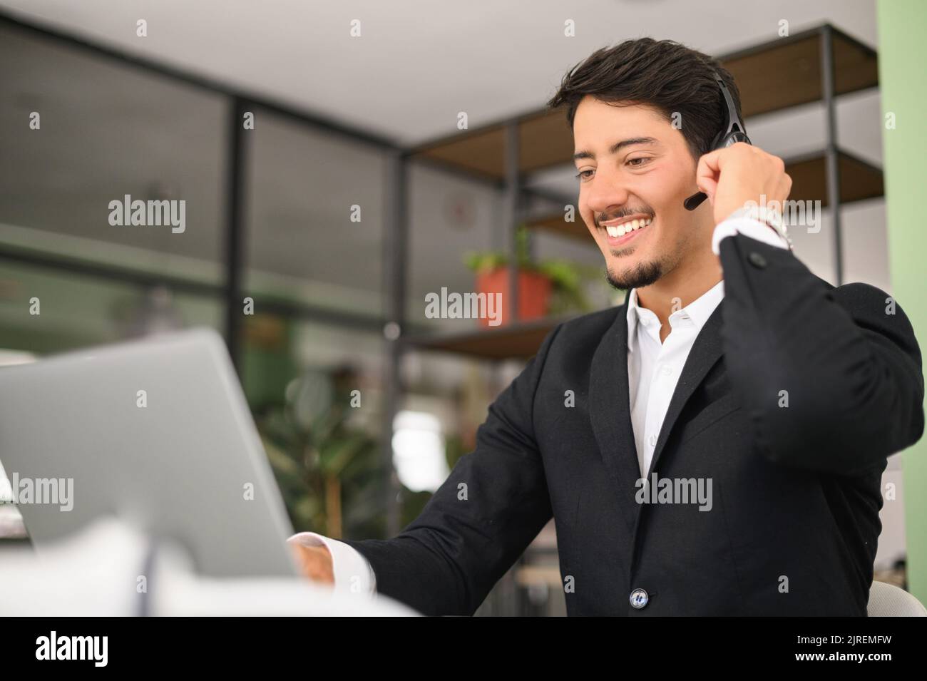 Ritratto di uomo latino fiducioso in cuffie wireless utilizzando computer portatile seduto in uno spazio ufficio contemporaneo, un dipendente maschio sta effettuando una videochiamata, parlando online a distanza Foto Stock