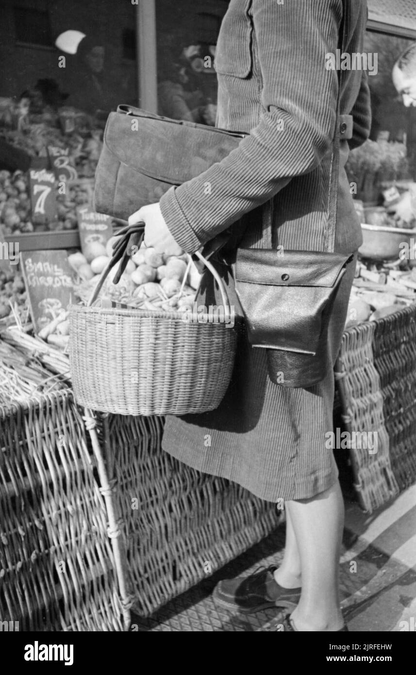 Una casalinga che trasportano Shopping basket e maschera a gas contenitore negozi presso un fruttivendolo a Londra durante il 1941. Una vista ravvicinata della signora giornata di shopping basket e maschera a gas caso che lei ha preso con lei per il suo viaggio di shopping. La maschera a gas caso ha una tasca speciale che gli consente di essere utilizzato come una borsetta. Alle sue spalle il fruttivendolo organizza la sua mercanzia. Questa fotografia è stata probabilmente preso sul King's Road nel quartiere di Chelsea. Foto Stock