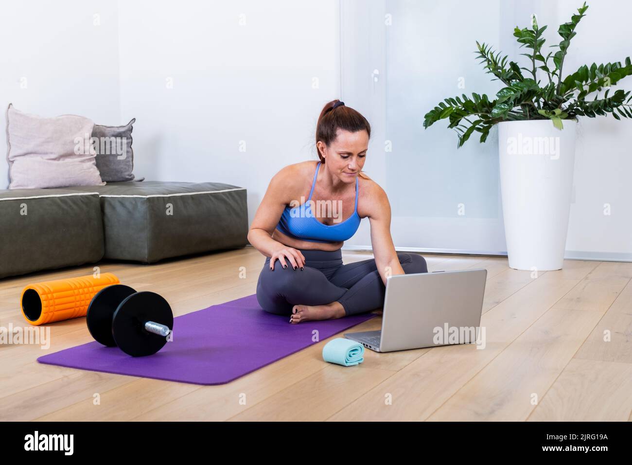Adatta la giovane donna in abbigliamento sportivo alla ricerca di corsi di formazione video sul web utilizzando un laptop, sdraiato sul tappetino yoga nel moderno soggiorno Foto Stock